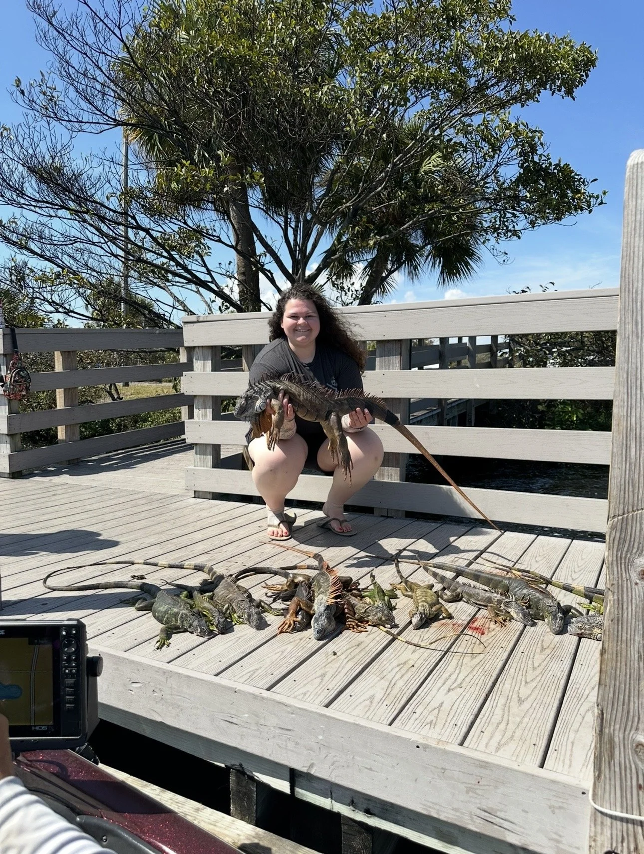 A woman crouching on a wooden deck holding a large fish, with several other fish laid out on the deck in front of her. There is a tree and blue sky in the background.