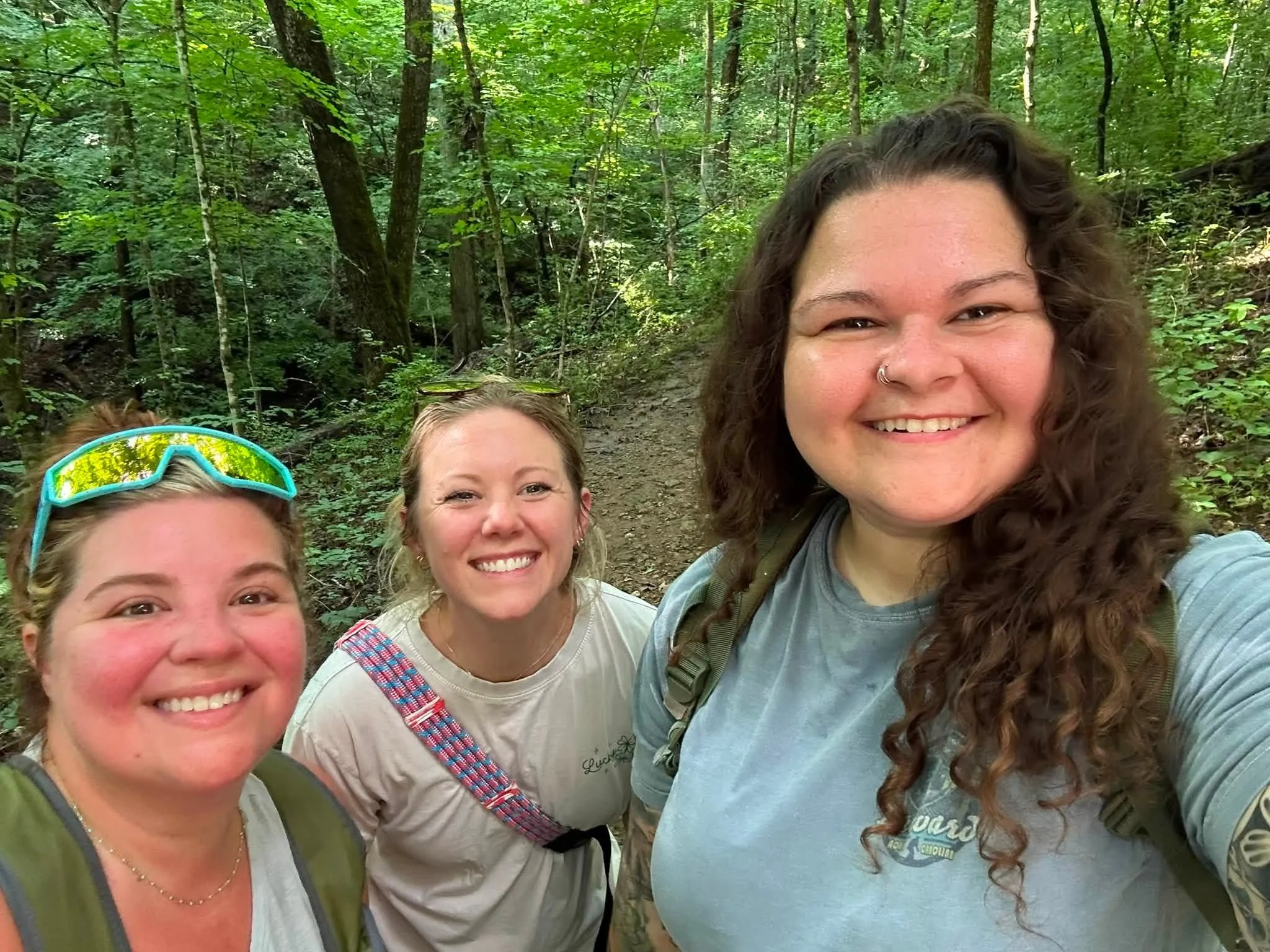 Smiling group of four women standing on a wooded trail during daytime, surrounded by green trees.