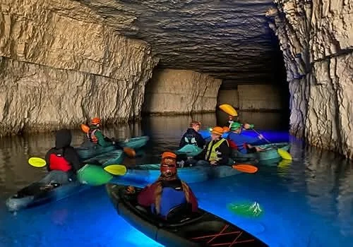 Group of people kayaking inside a cavern with rocky walls and a small opening at the back.