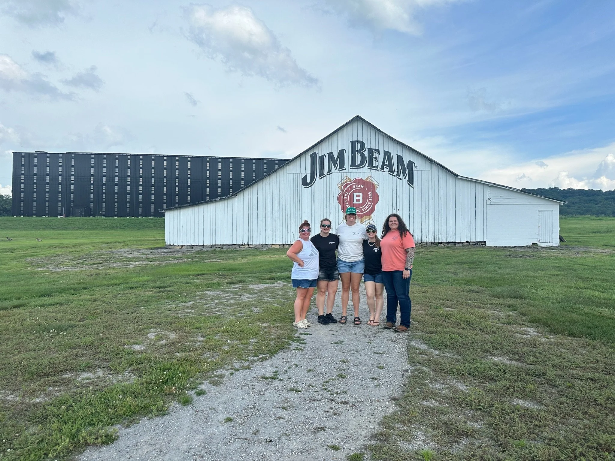 A group of five women standing together outside in front of a white barn with the words "Jim Beam" and a red bourbon whiskey logo on it. There is a large, modern dark building in the background, a cloudy sky, and green grass and dirt underfoot.