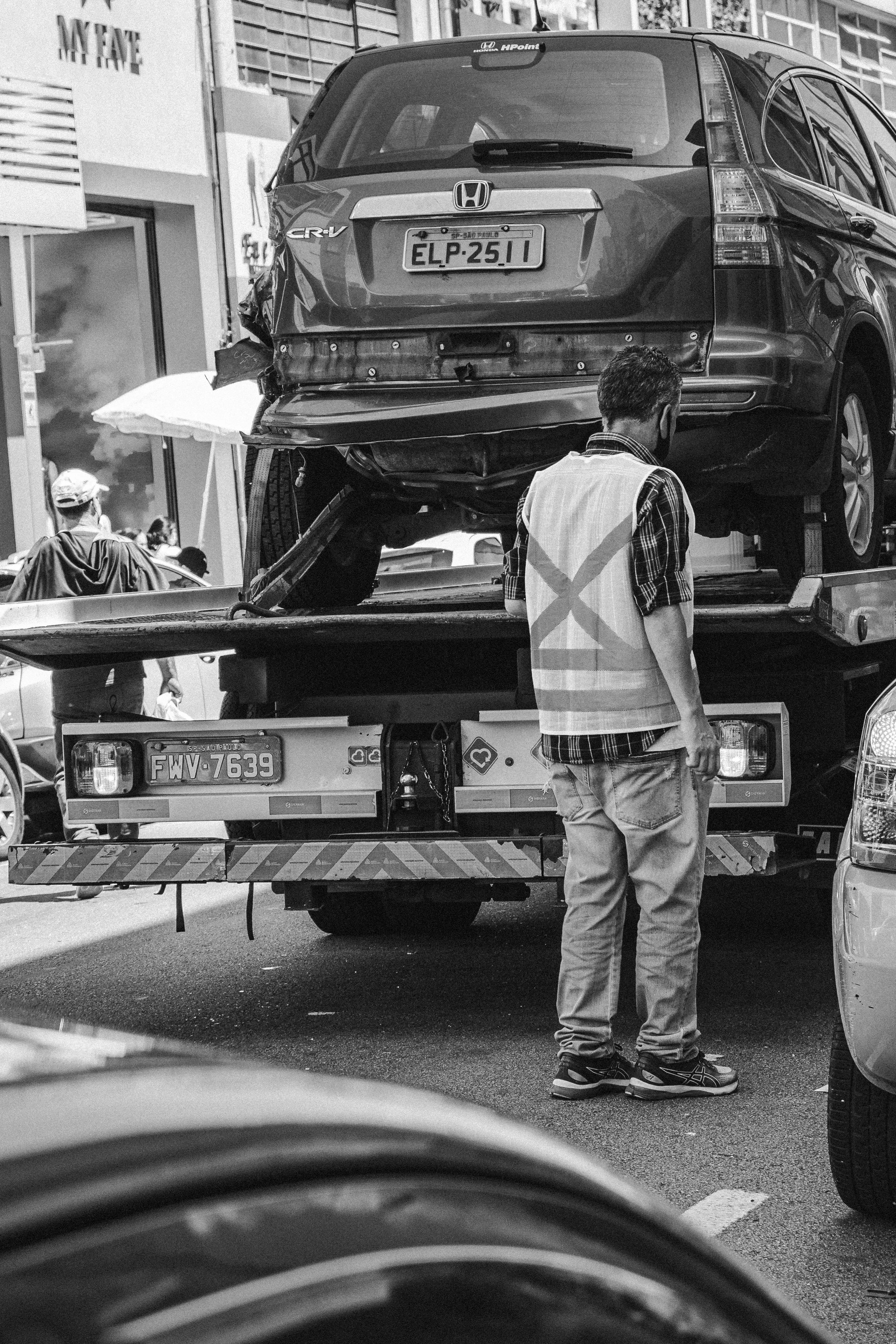 Rear view of a damaged Honda CR-V being transported on a flatbed tow truck, with a man wearing a checkered shirt, jeans, and a mask standing next to it in an urban area.