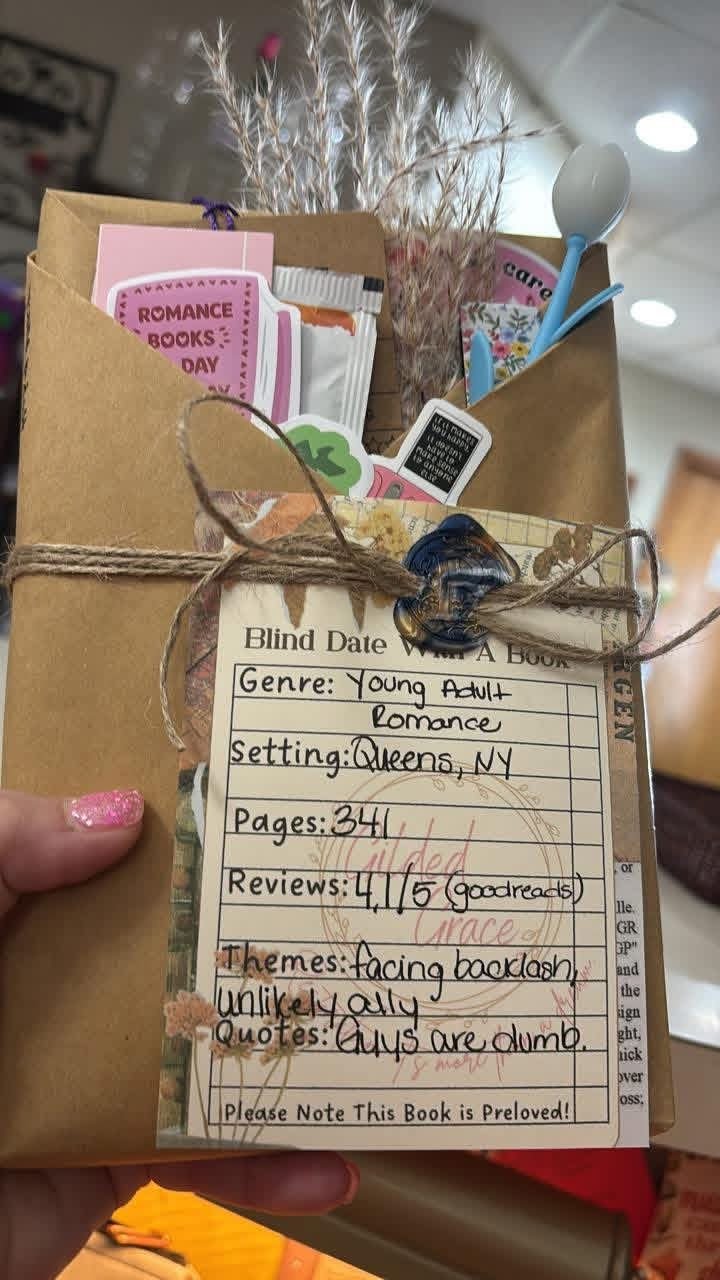 A gift wrapped in brown paper with a tag that resembles a book review form, containing details about a book titled 'Blind Date with a Book'. The package is decorated with dried flowers, a ribbon, and a wax seal. Inside are small gift items like a tiny spoon, snack pack, and colorful bookmarks.