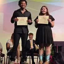 Two students holding certificates on stage at an awards ceremony, with two people sitting in the background.