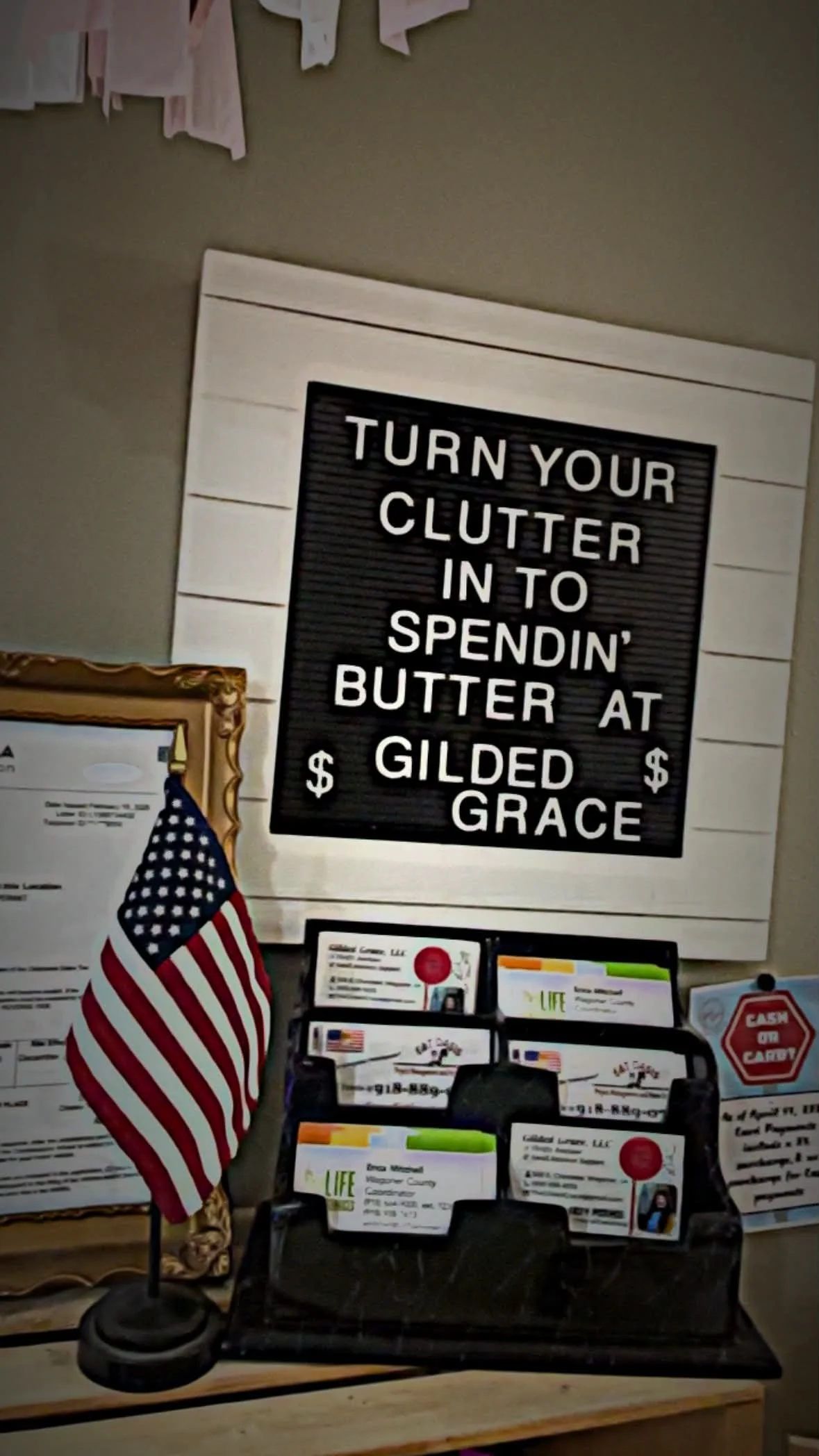 A black letter board with white letters that says, "Turn your clutter into spendin' butter at Gilded Grace." A small American flag on a stand is in front of the board, and there are various brochures and papers on a rack below it.