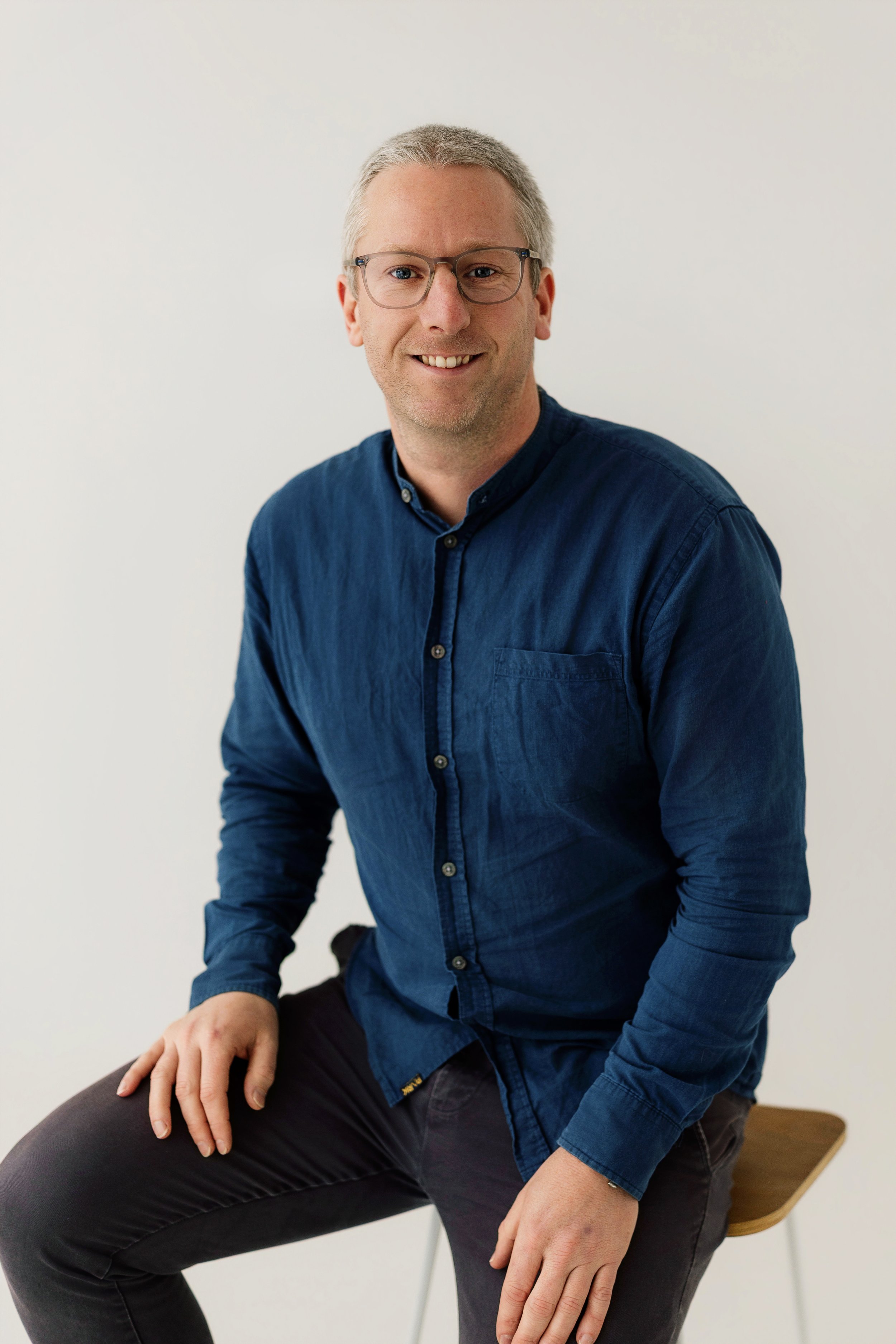 A man with glasses and gray hair sitting on a wooden chair against a plain white wall. He is wearing a dark blue button-up shirt and black pants, smiling at the camera.