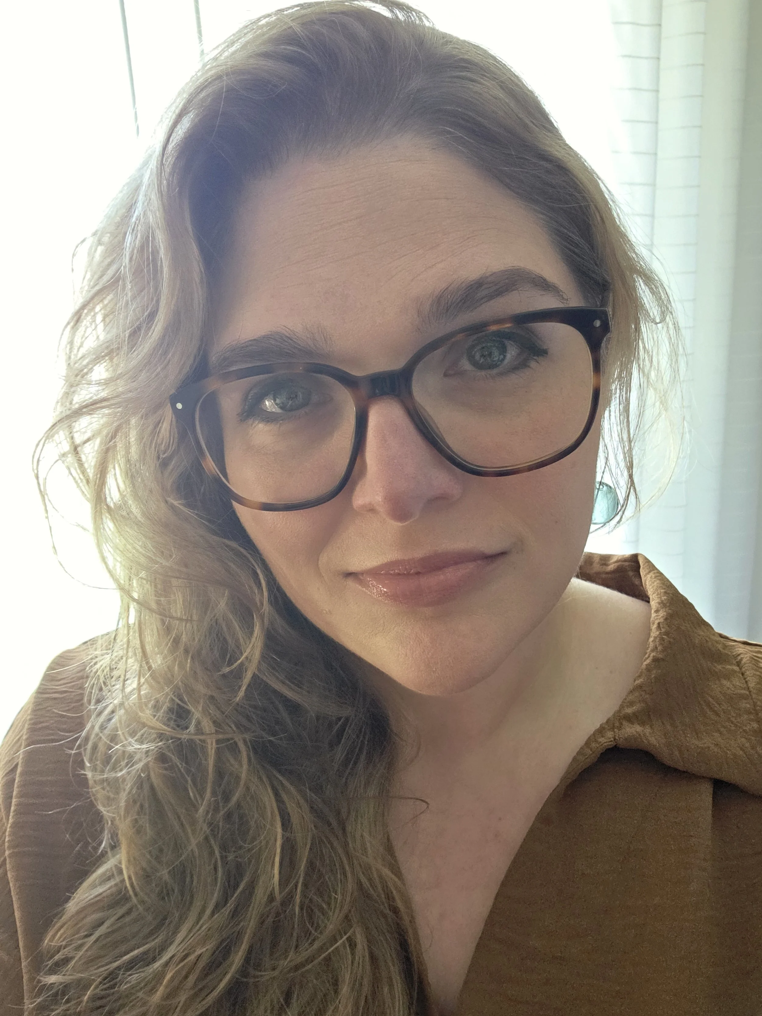 A woman with curly blonde hair wearing black-rimmed glasses and a brown top, sitting near a window.