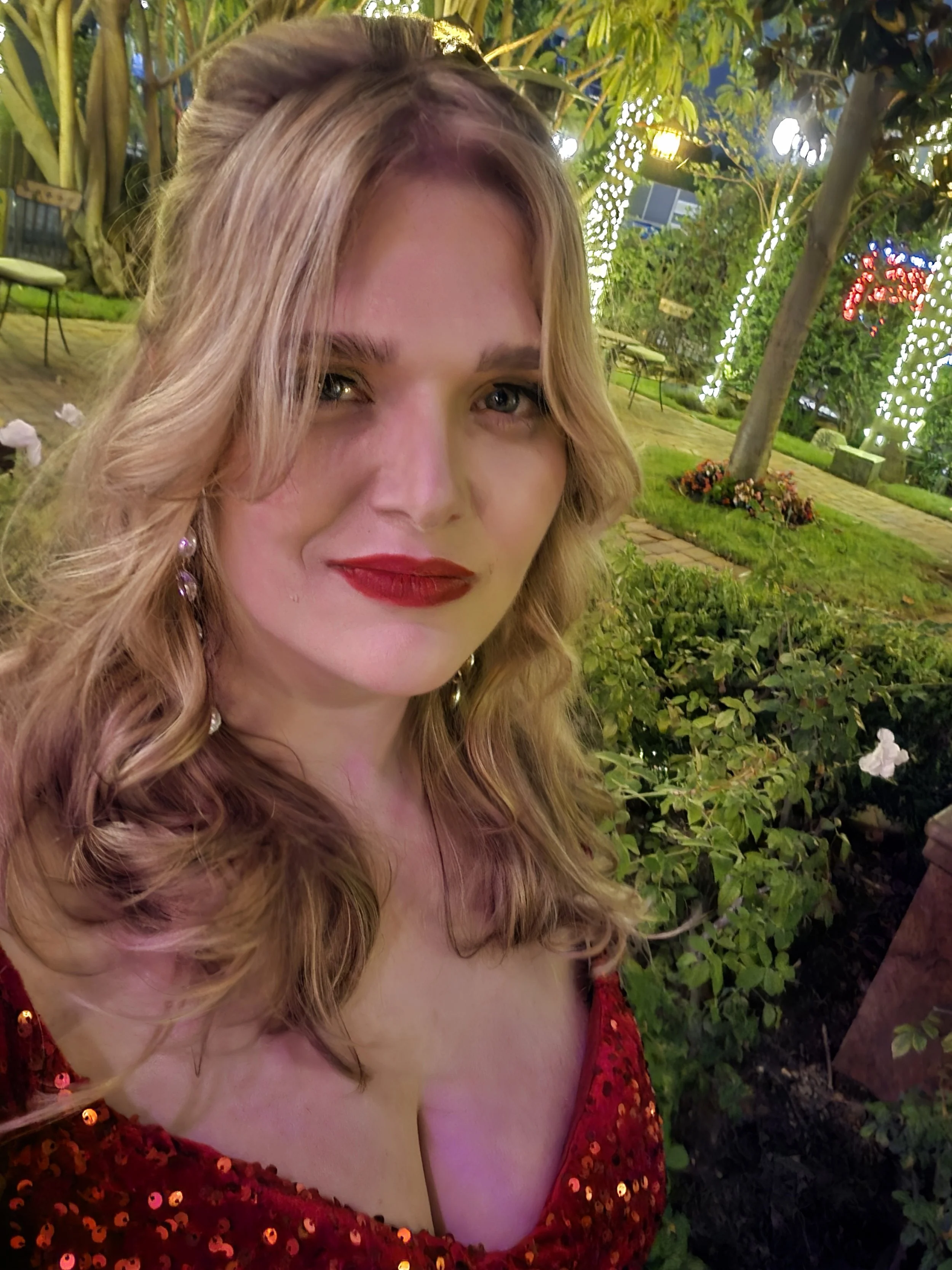 A woman in a red, sequin dress with blonde hair and red lipstick at a decorated outdoor event at night.
