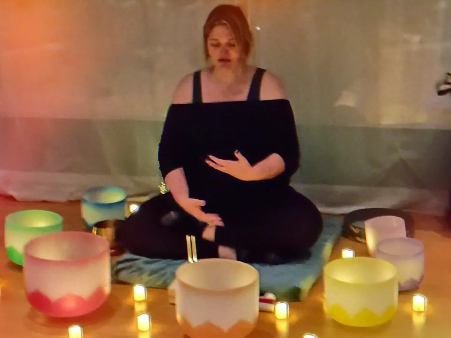 A woman sitting cross-legged on a cushion surrounded by colorful singing bowls and small candles in a dimly lit room.