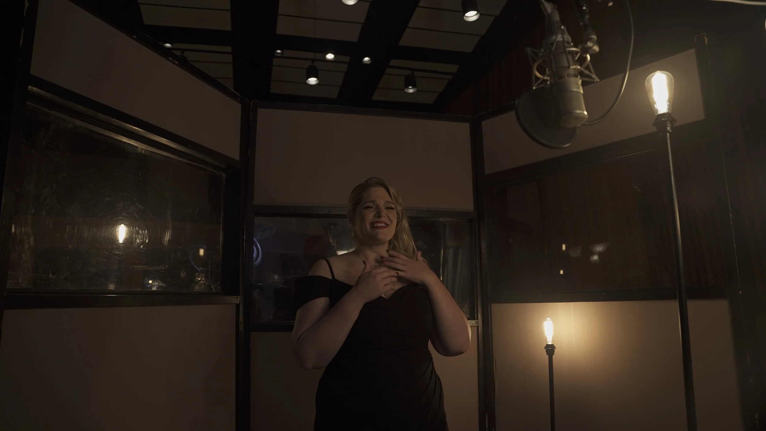 A woman in a black evening dress smiling and touching her chest, standing in a dimly lit room with vintage-style exposed light bulbs and dark wood paneling.