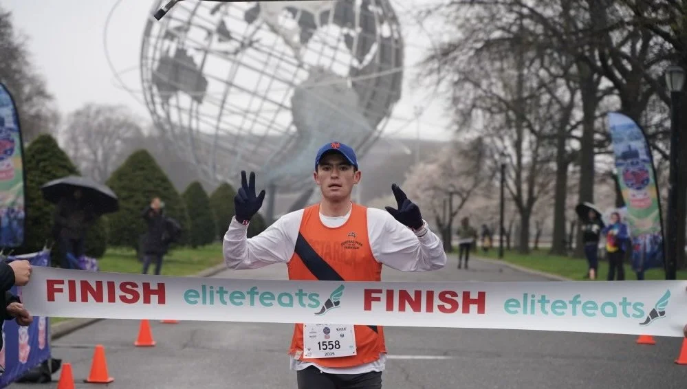 A young man in athletic gear crossing the finish line of a race in front of the UN's Queen Elizabeth II Great Globe sculpture, with trees and some spectators in the background, and a banner reading 'FINISH elitefeats'.