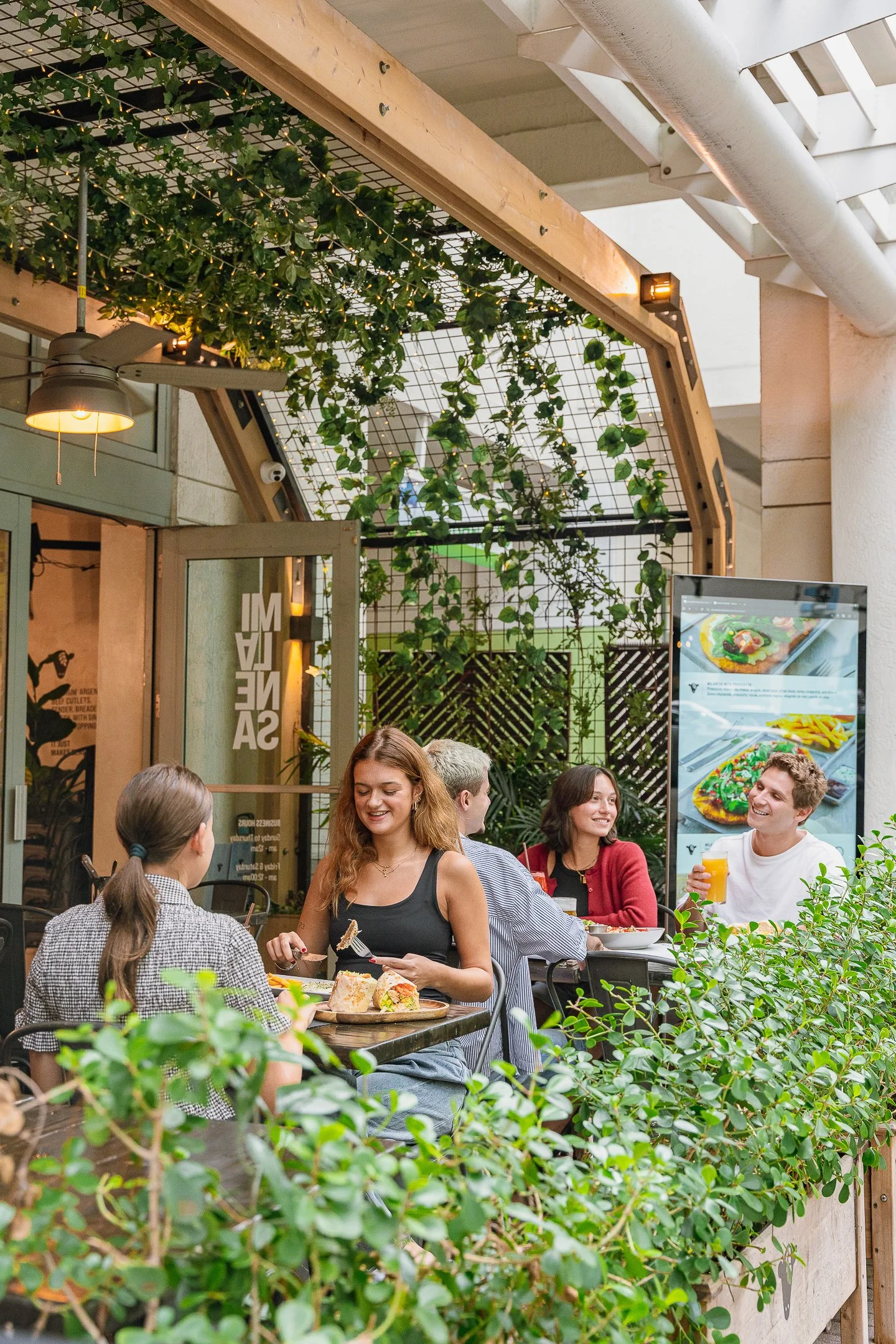 Grupo de personas comiendo y conversando en un patio de restaurante con plantas y decoración moderna.