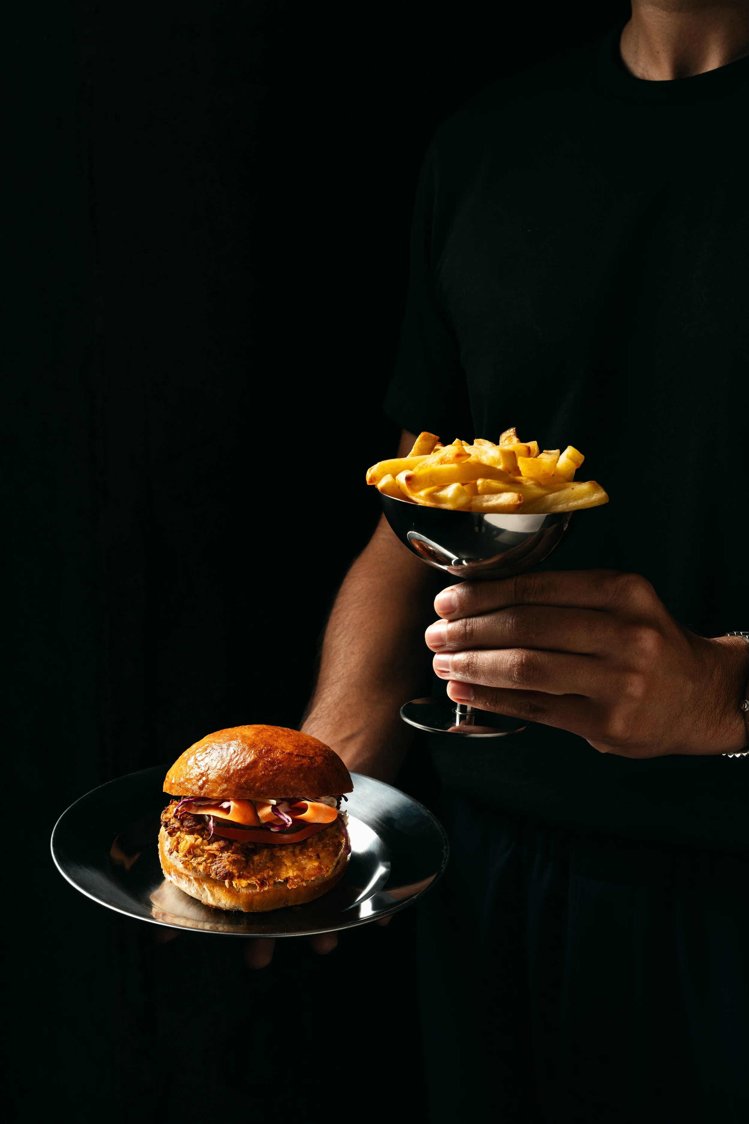 Persona sosteniendo un plato con papas fritas y un plato con una hamburguesa en un fondo oscuro