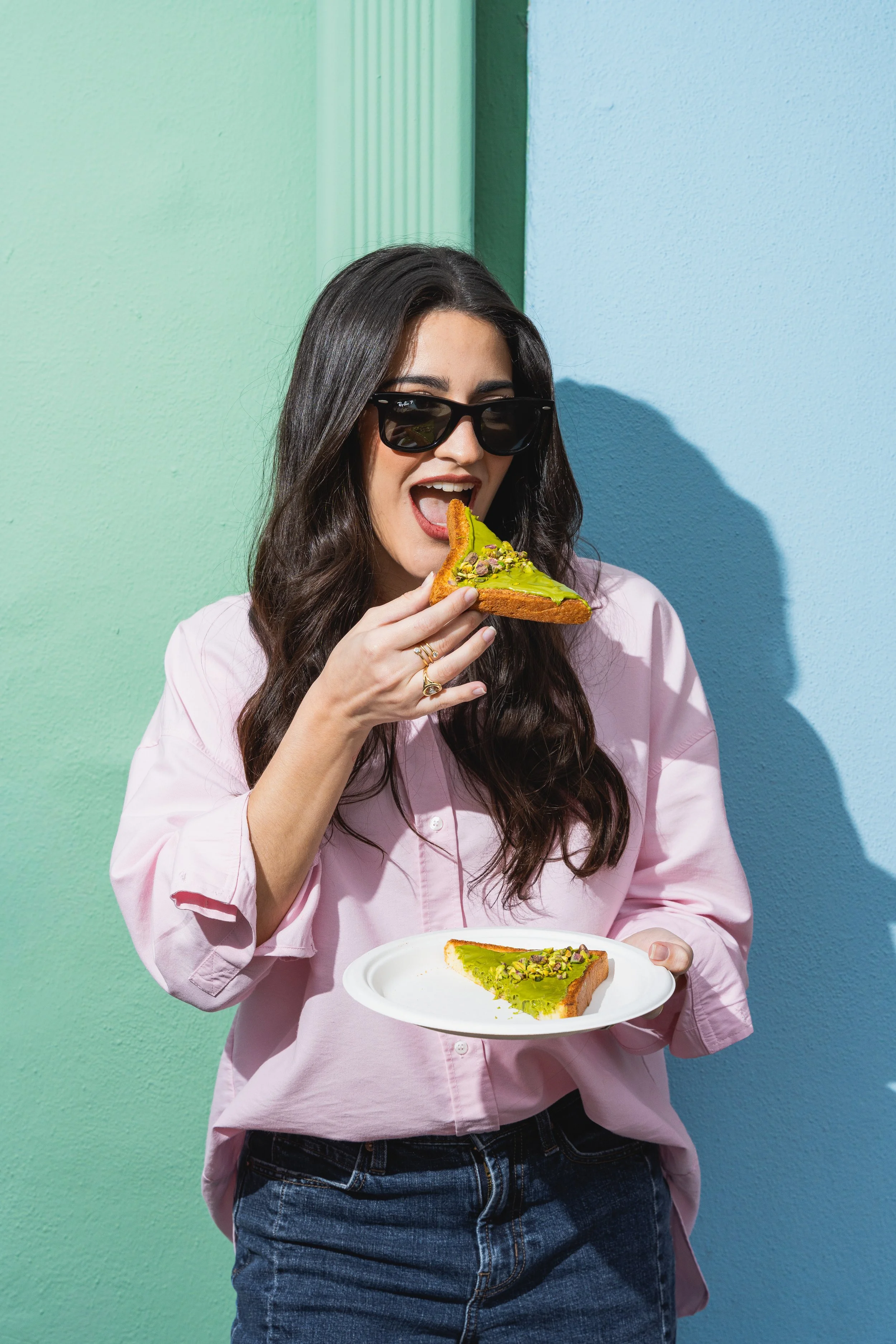 Mujer con gafas oscuras sosteniendo una rebanada de pan con aguacate y pistachos, y una placa con otra rebanada de pan con aguacate, en un fondo de pared azul y verde.