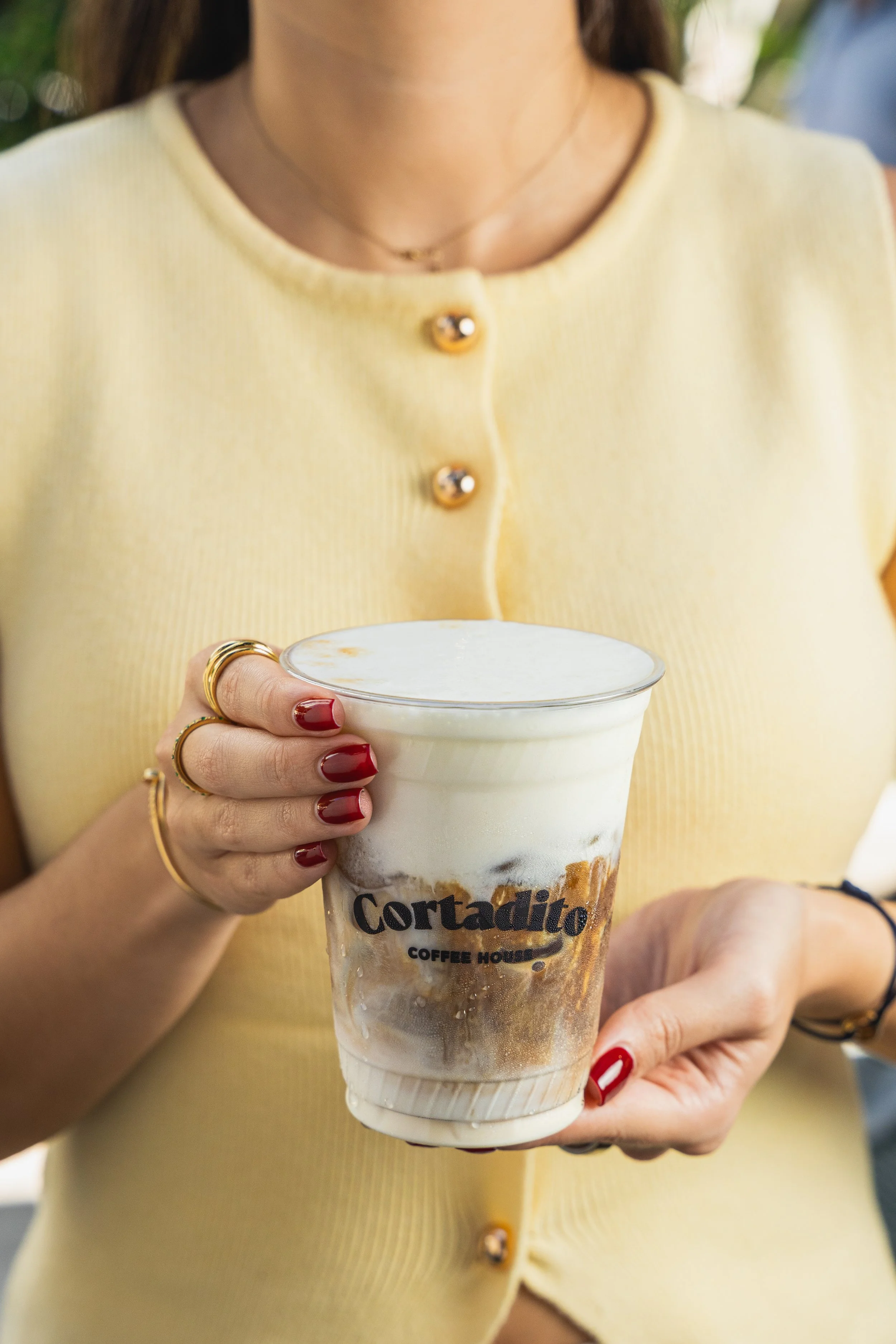 Persona sosteniendo un vaso de café con leche y caramelos en un lugar exterior, con un fondo de vegetación.