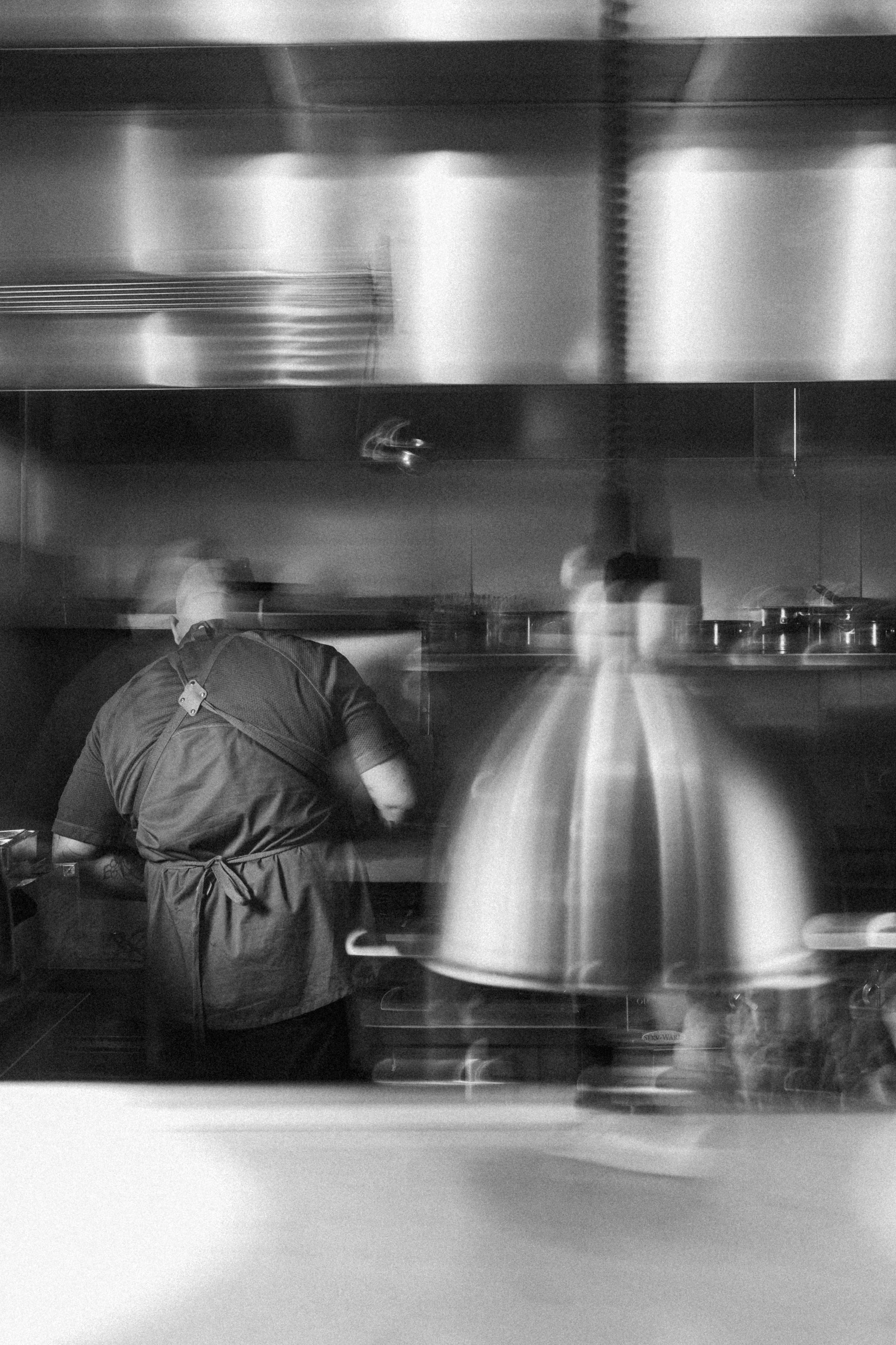 Una imagen en blanco y negro de una cocina de restaurante. Hay un chef de espaldas en primer plano, vestimenta de cocina y un delantal. Al fondo, hay una estufa con ollas y utensilios, y un caballero parece estar preparando comida. La escena tiene movimiento difuso por la velocidad de la cámara.