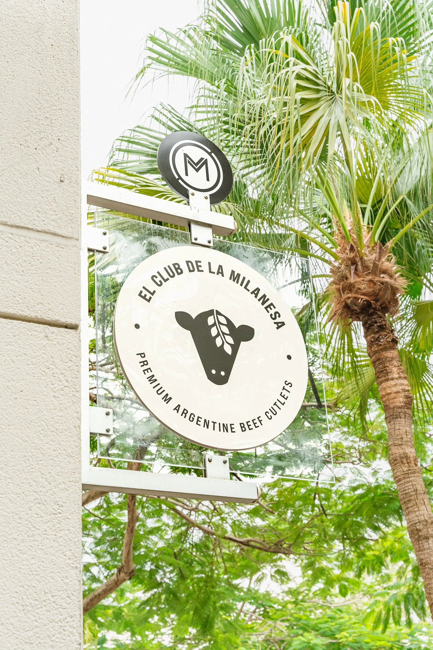 Se observa un cartel de un club de carne de res argentino de altísima calidad en un entorno con palmeras verdes y cielo brillante.