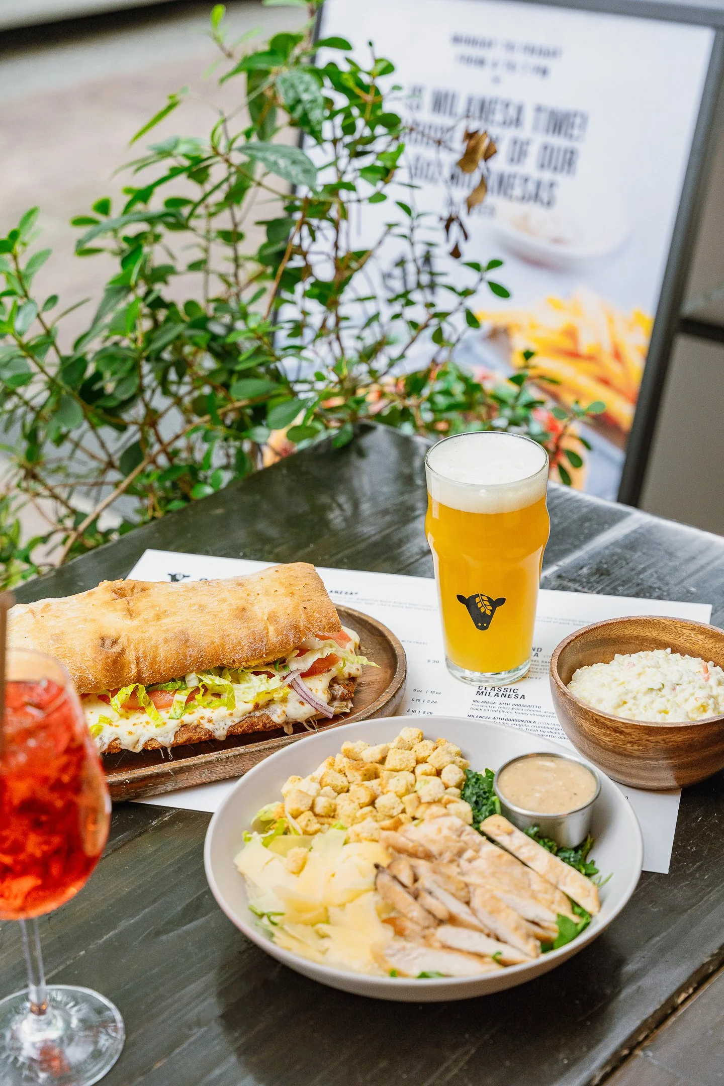 Plato con ensalada, un sándwich y acompañamientos, en una mesa de madera, con una bebida en un vaso y una planta decorativa en el fondo.