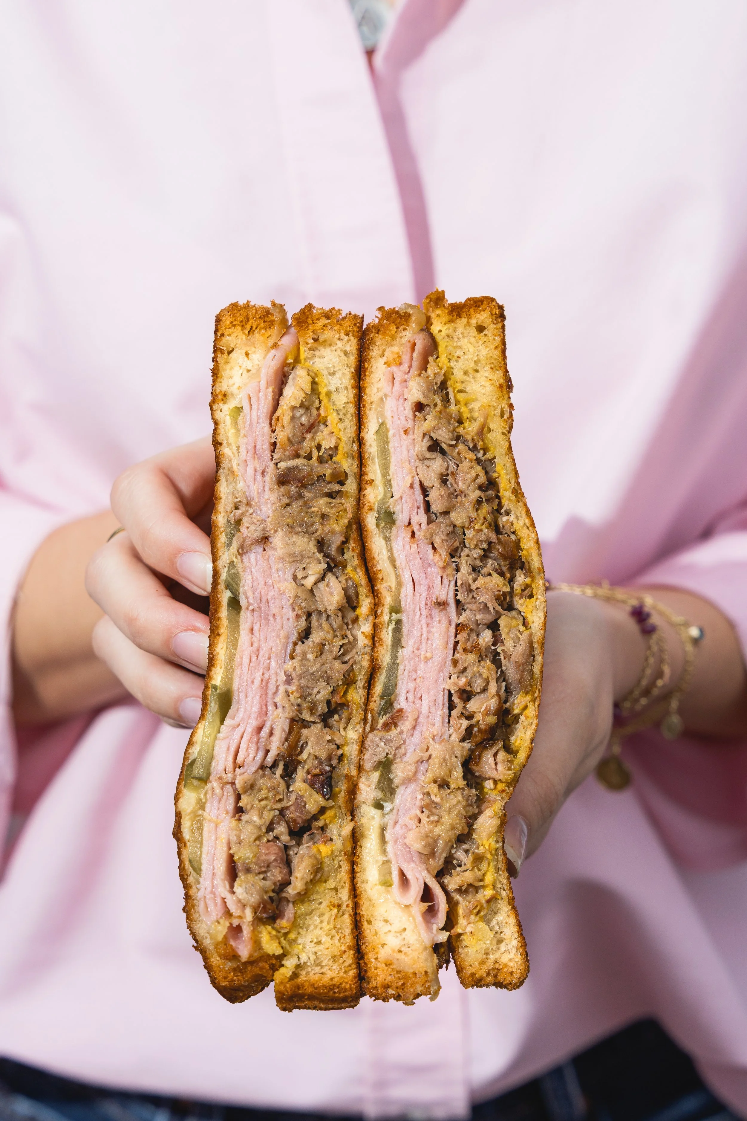 Persona sosteniendo un sándwich de carne en capas con pan dorado y relleno de carne, jamón, queso, pepinos y vegetales, con fondo rosa.