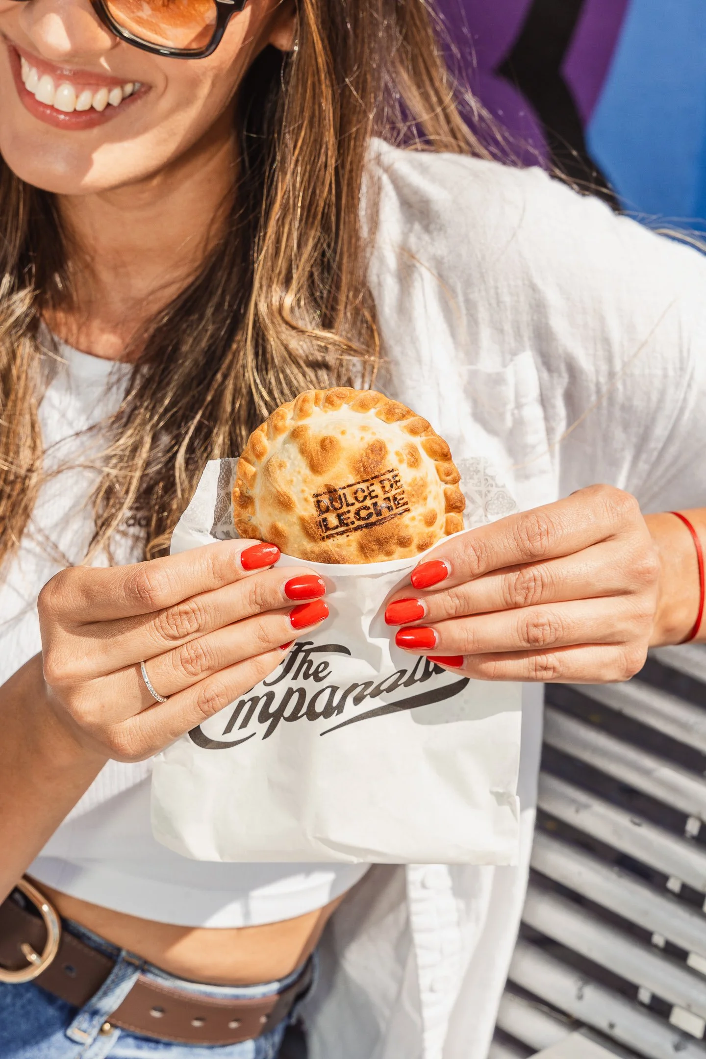 Mujer sonriendo sostiene una empanada con la marca 'DULCE DE LECHE' en la parte superior, lleva uñas rojas, lleva gafas y viste una chaqueta blanca, con un fondo de banco de madera.