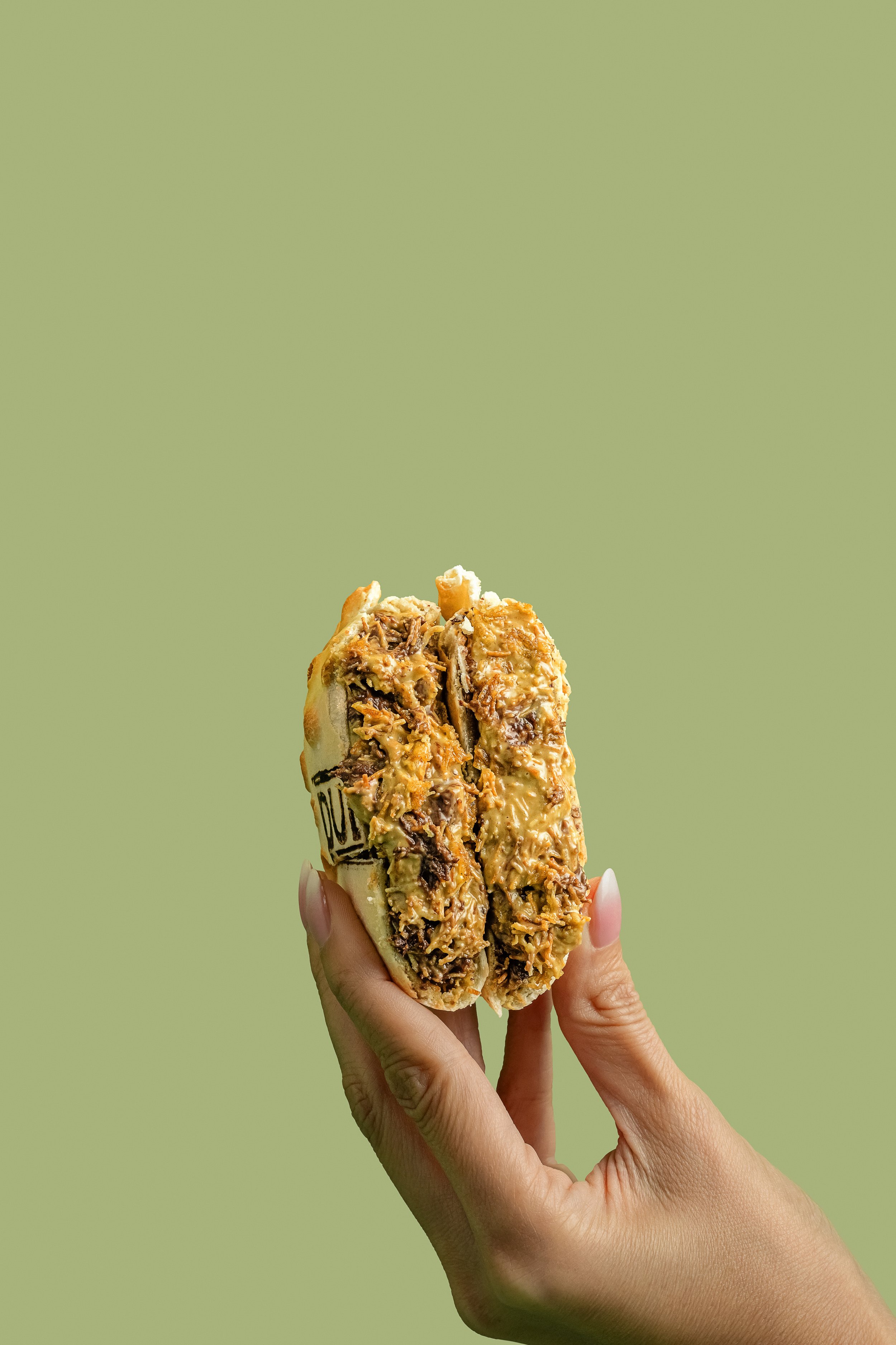 Una mano sostiene un sándwich abierto con relleno de carne, queso y vegetales, contra fondo de color verde claro.