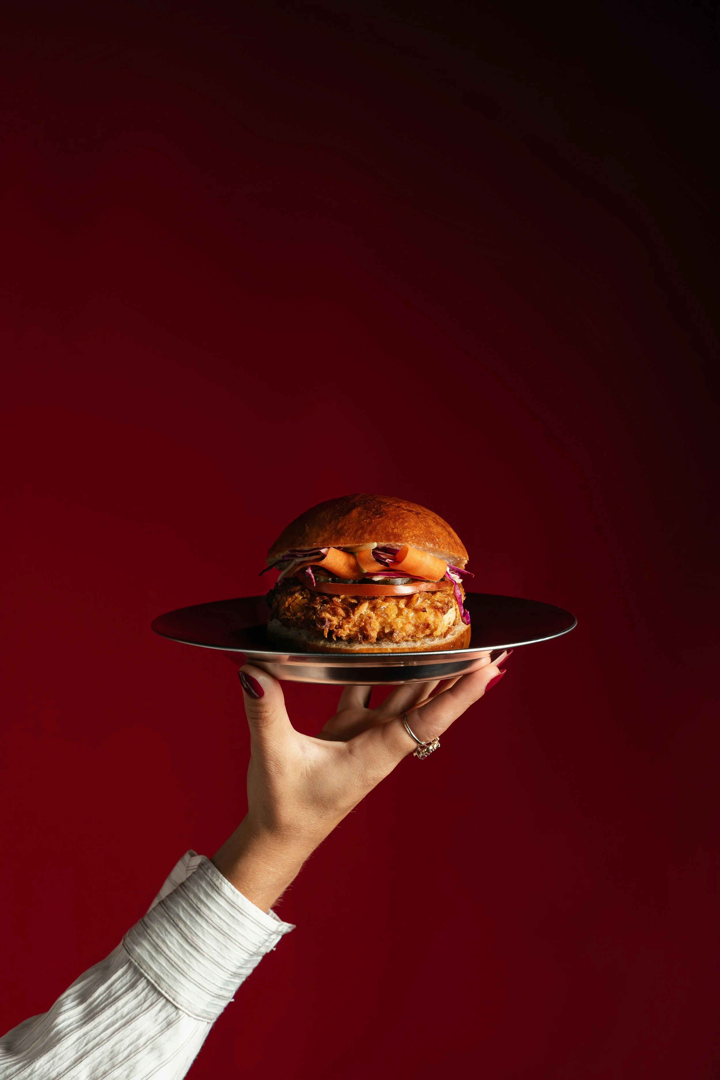 Una mano con uñas pintadas de rojo sostiene un plato negro con una hamburguesa con pan, carne, queso, vegetales y salsas, en fondo rojo oscuro.