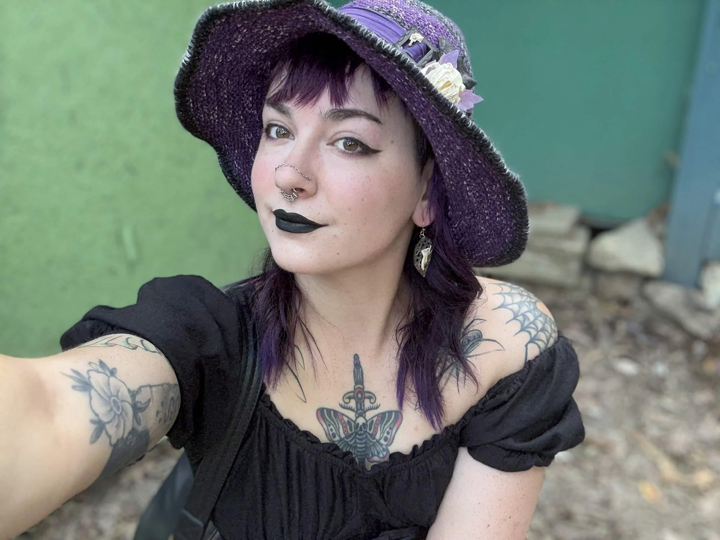 A woman with short purple hair and tattoos on her arms and chest, wearing a large purple hat with decorative flowers and a middle part, taking a selfie outdoors with a green and blue background.