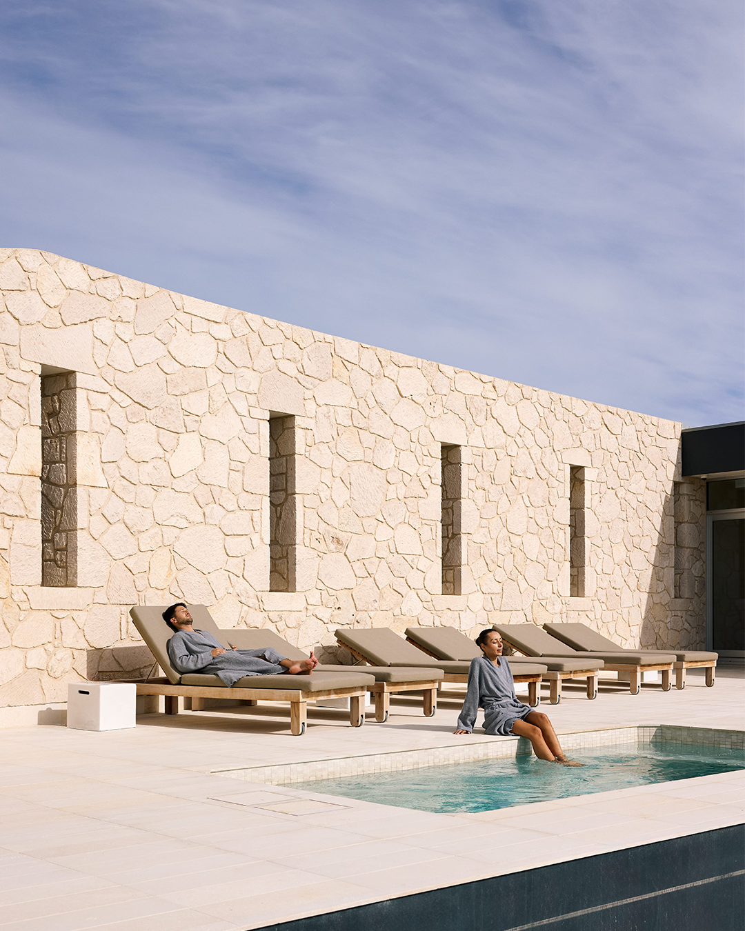 People relaxing by a pool on lounge chairs, with a woman dipping her feet in the water, against a stone wall and cloudy sky.