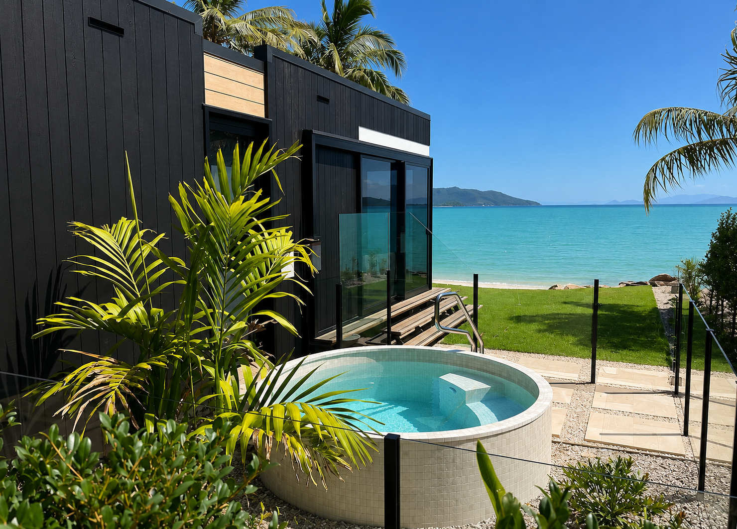 A modern house overlooking a beach with a tiled plunge pool in the backyard surrounded by tropical plants, with water and distant land visible in the background.
