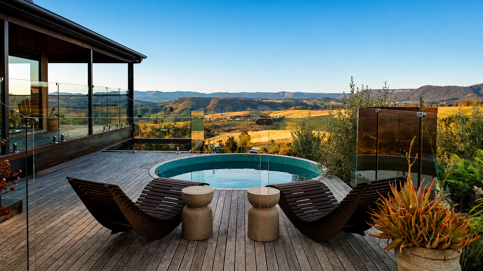 A rooftop deck with a small round pool, two lounge chairs, and a scenic view of rolling green hills and mountains in the background during daytime.