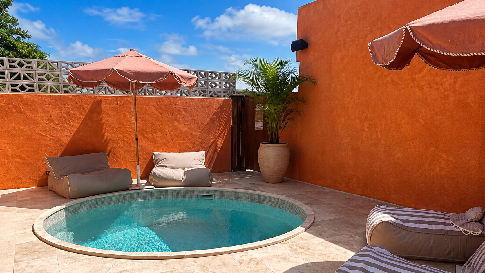 A small round swimming pool with seating cushions and bean bags on a tiled patio, orange walls, a large potted palm, and umbrellas providing shade, under a partly cloudy blue sky.
