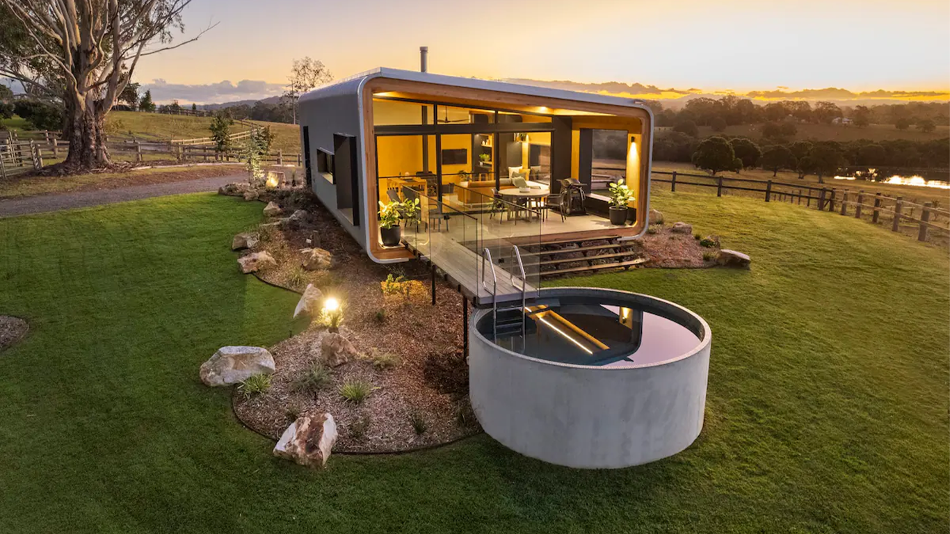 Modern tiny house with large windows, outdoor deck, and reflecting pool in a rural landscape during sunset.