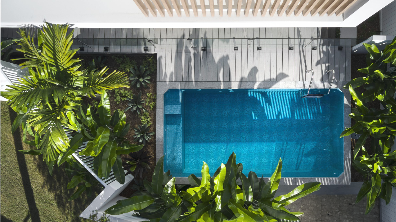 View from above of a small rectangular swimming pool surrounded by lush green plants and trees, with a deck area and glass railing.