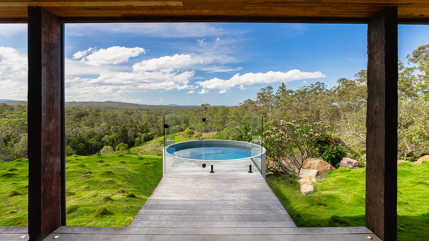 View of a small, round pool at the end of a wooden deck, with a glass fence, overlooking a lush green landscape with trees and a partly cloudy blue sky.