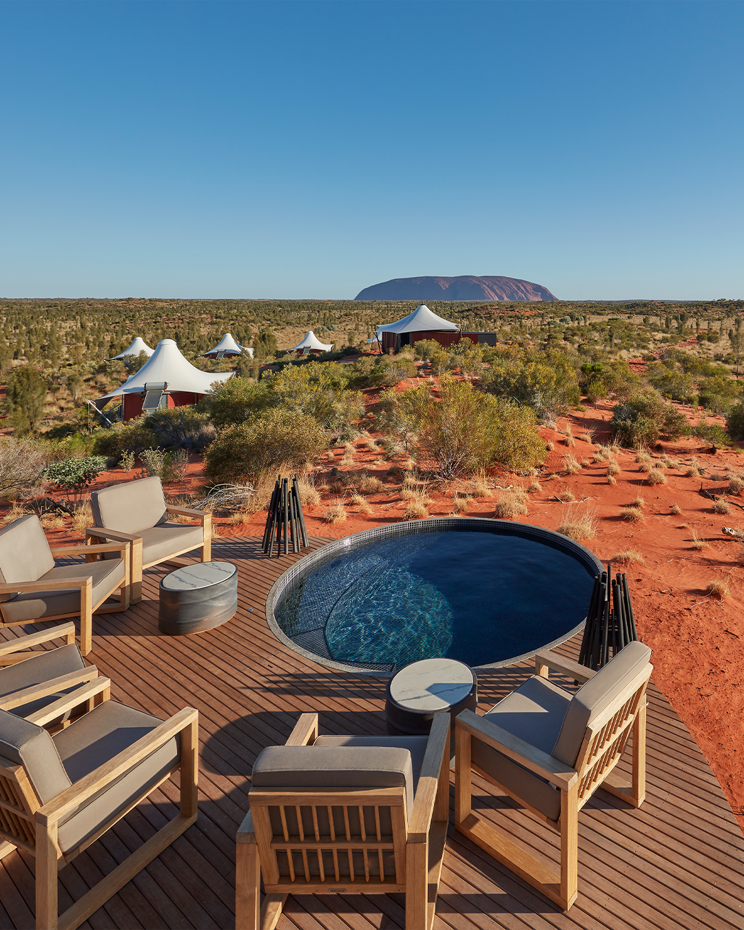 Outdoor living space with wooden deck, chairs, and small round table surrounding a hot tub, set in a desert landscape with red sand, sparse vegetation, and distant dome-shaped tents and a mountain in the background under a clear blue sky.
