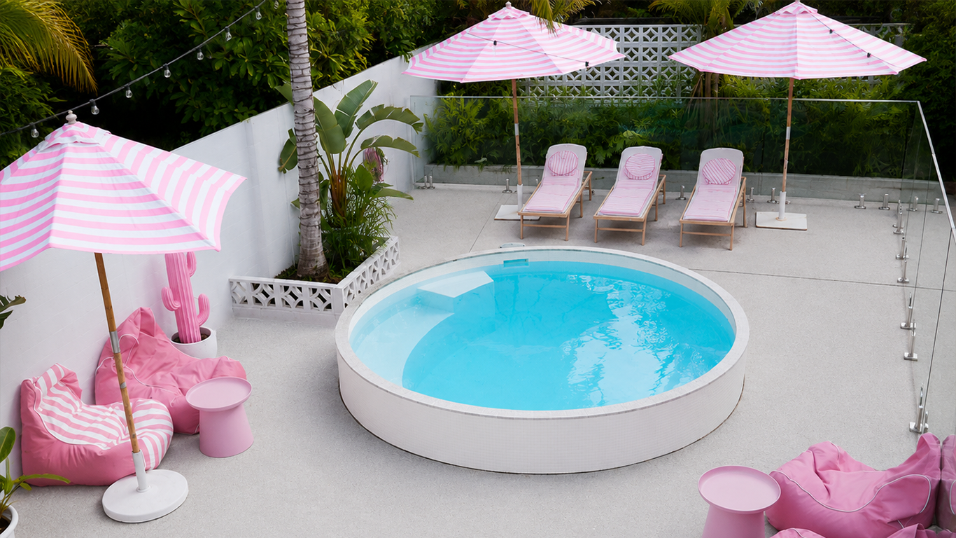 An outdoor pool area with pink and white striped umbrellas, pink bean bags, and lounge chairs with pink towels, surrounded by green plants and a white fence.