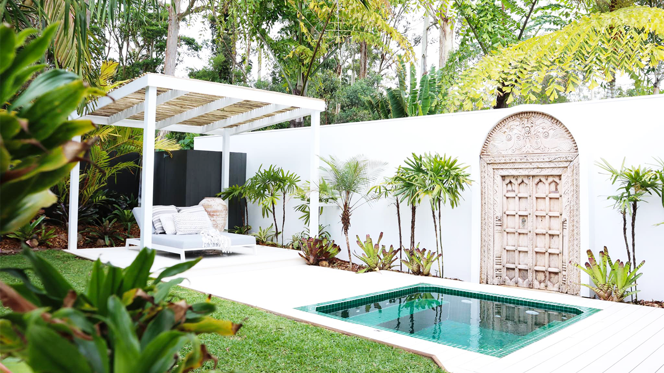 Backyard with a small pool, white lounging bed with pillows under a white pergola, surrounded by green plants and trees, decorative stone door on white wall.