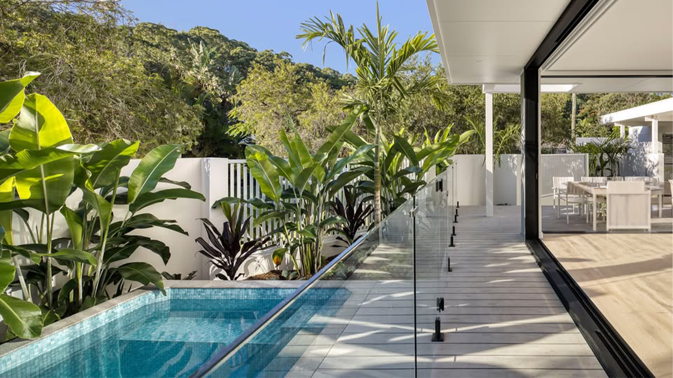 Modern balcony with glass railing overlooking lush green trees and hills, with a small pool and outdoor dining area visible.