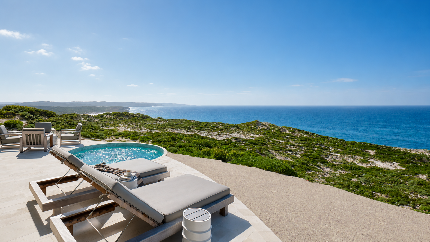 A porch with lounge chairs and a titled plunge pool overlooking a scenic ocean view with blue sky and green hillside.