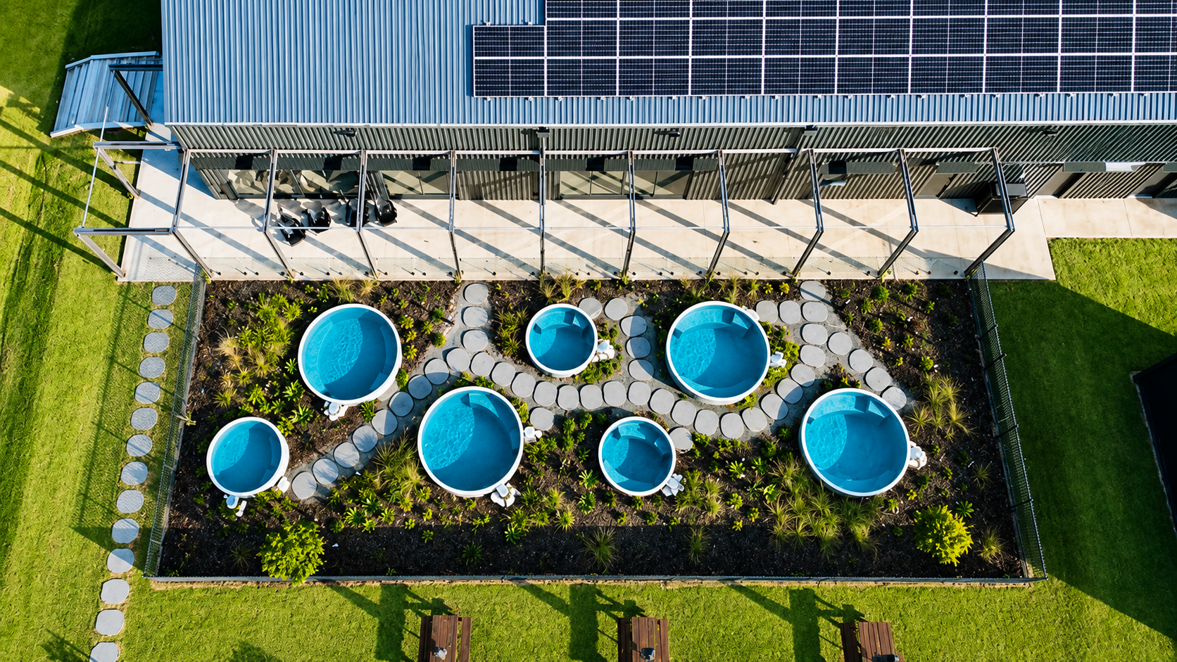 A rooftop garden with multiple round, blue hot tubs surrounded by lush greenery, a concrete path, and a modern building with solar panels.
