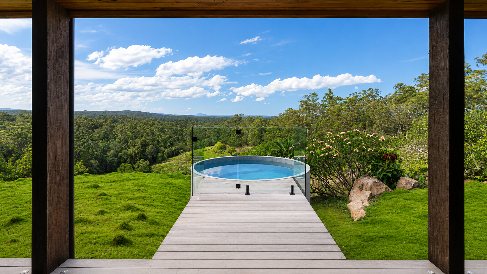 A tiled plunge pool on a wooden deck, with a glass safety barrier, overlooking a lush green landscape with trees, hills, and a blue sky with scattered clouds.