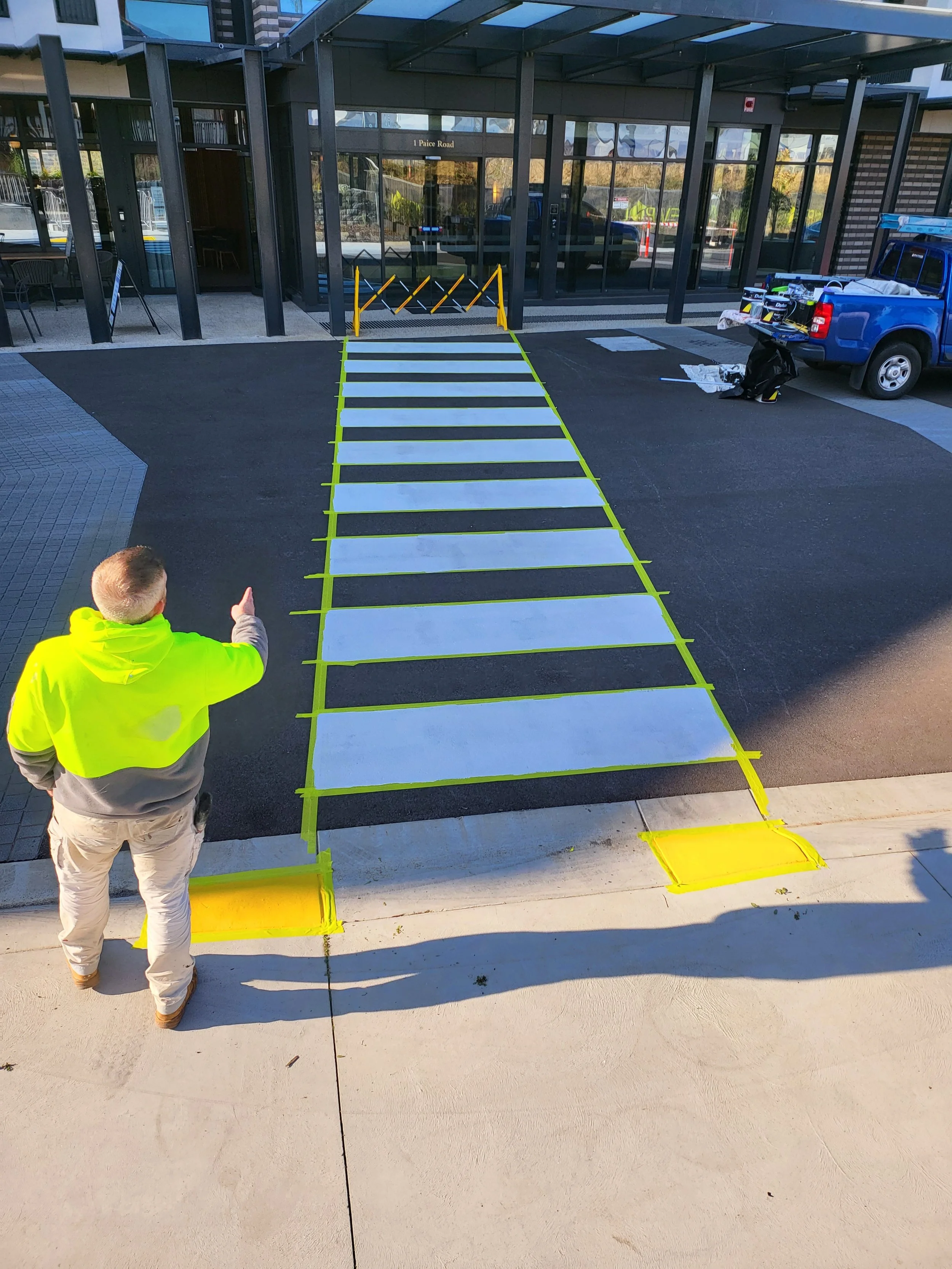 A person in a yellow jacket pointing at a newly painted crosswalk with white stripes and yellow borders in front of a glass building. There are construction materials and a blue truck nearby.