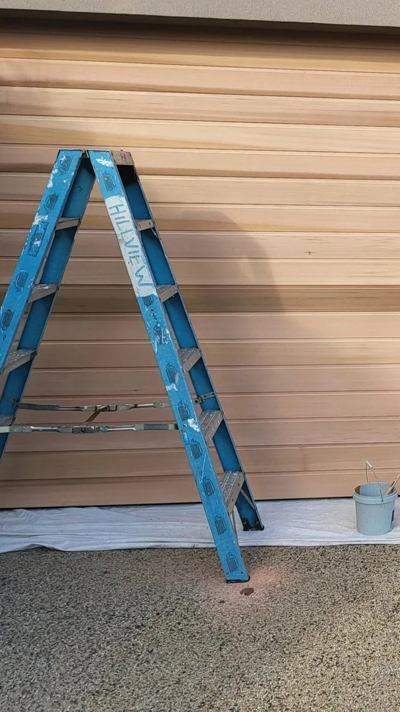 Blue ladder set up in front of a beige garage door, with a bucket of paint and a paintbrush on the floor nearby, indicating painting or maintenance work.