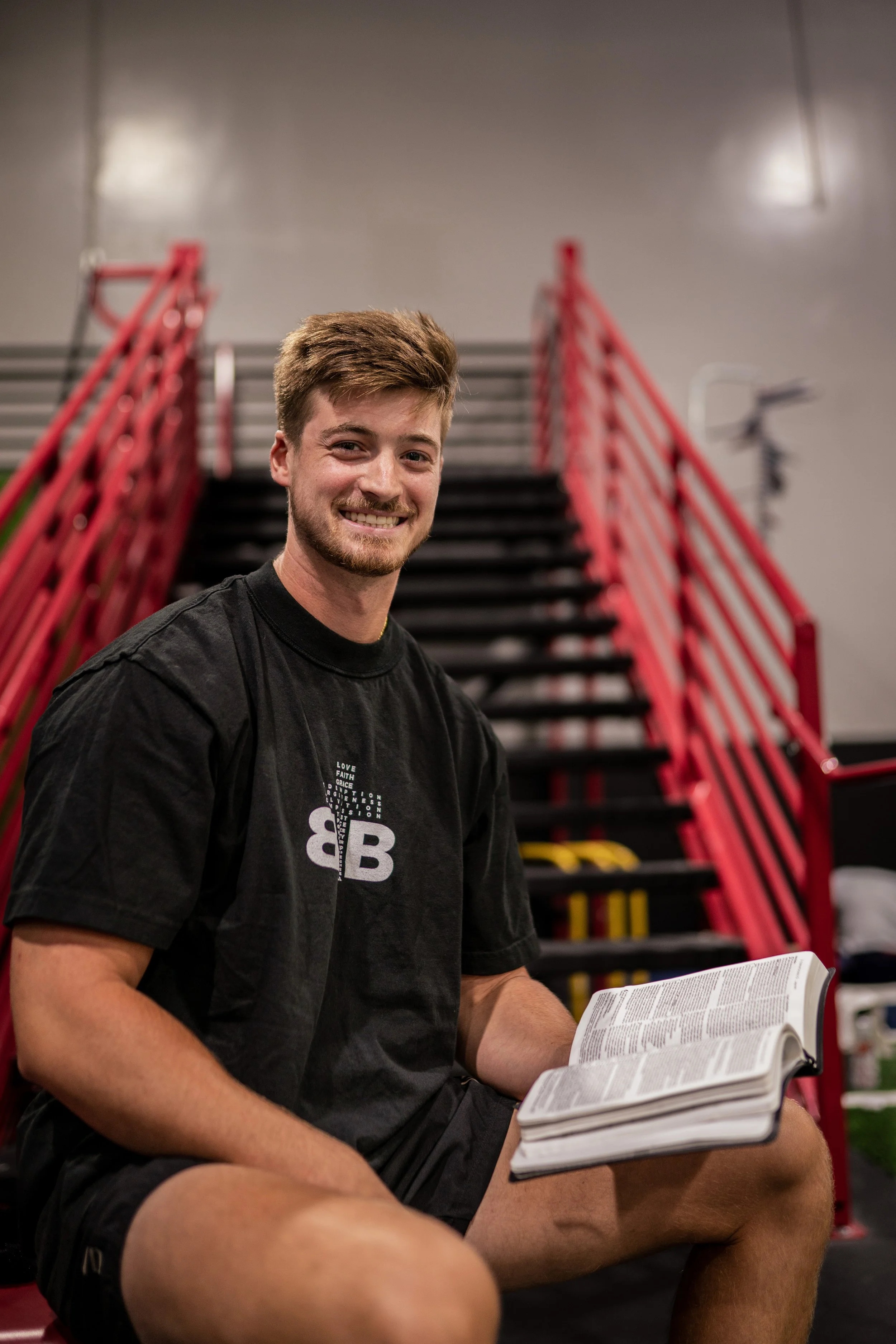 A young man with brown hair and a beard sitting on gym stairs, smiling, wearing a black t-shirt with white text and logo, holding an open book or magazine.