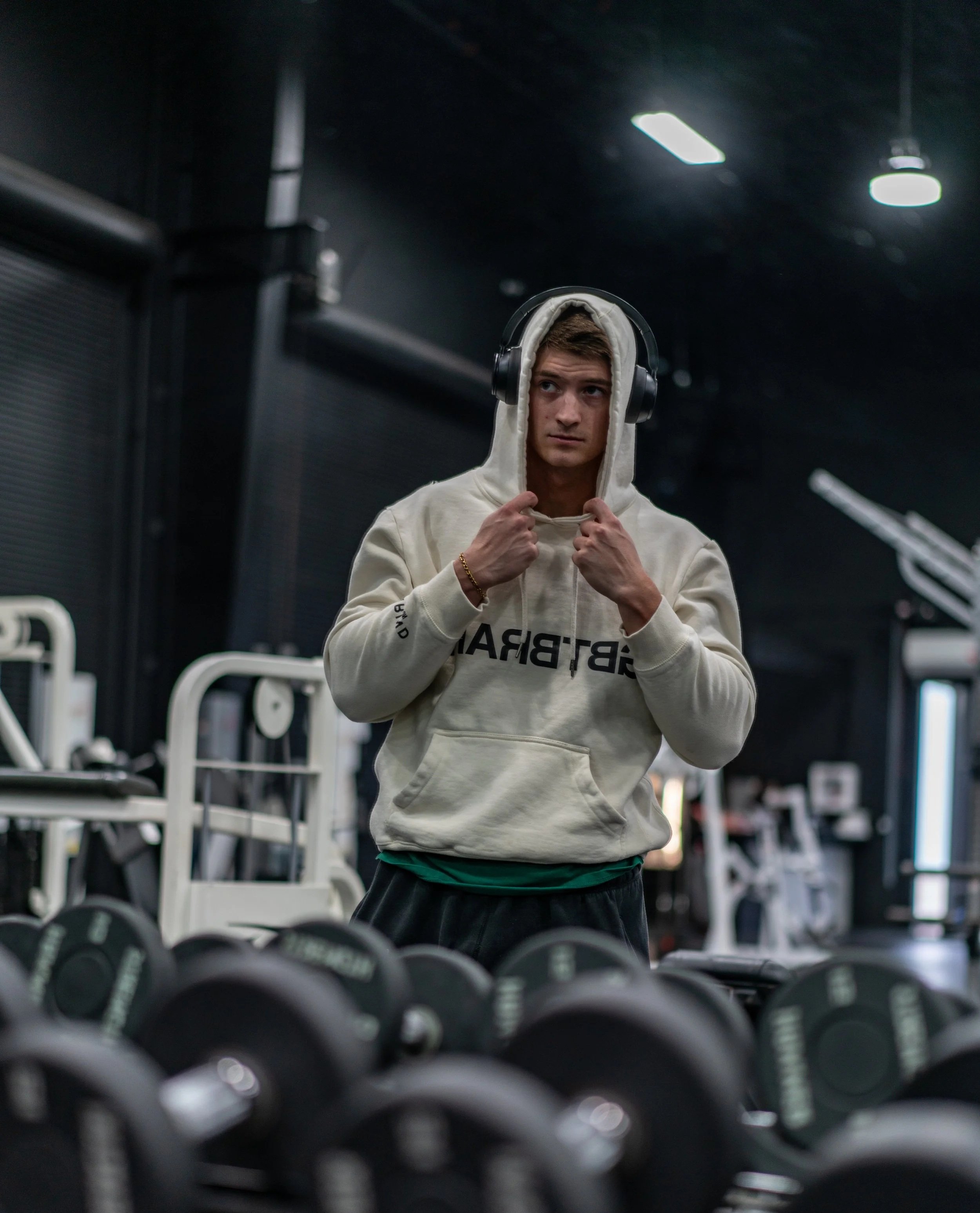 A young man wearing a hoodie and headphones standing in a gym surrounded by dumbbells.