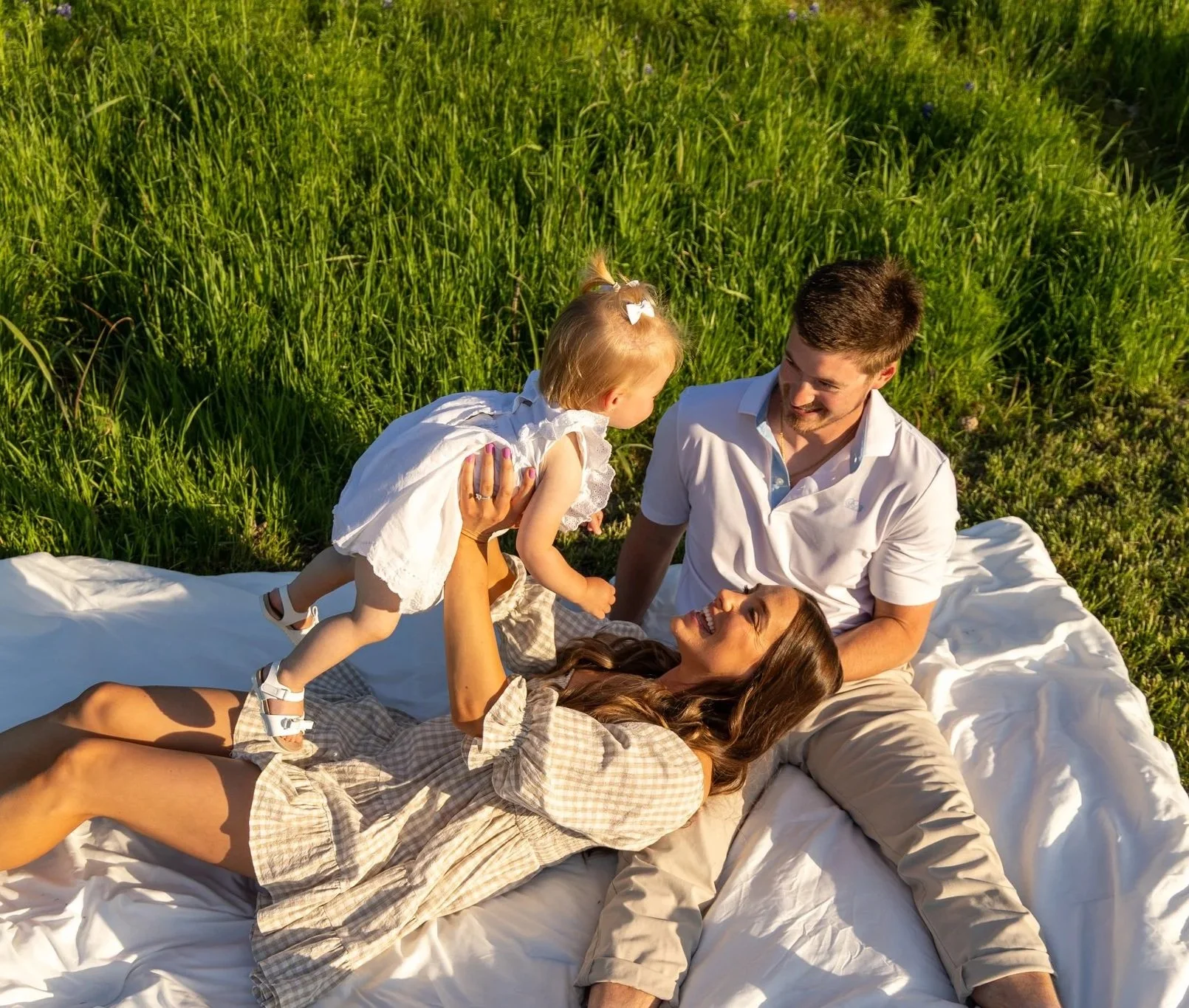 A family of four having a picnic on a blanket in a grassy field, with the mother lying down, the father sitting, and their young daughter standing and playing with her parents.