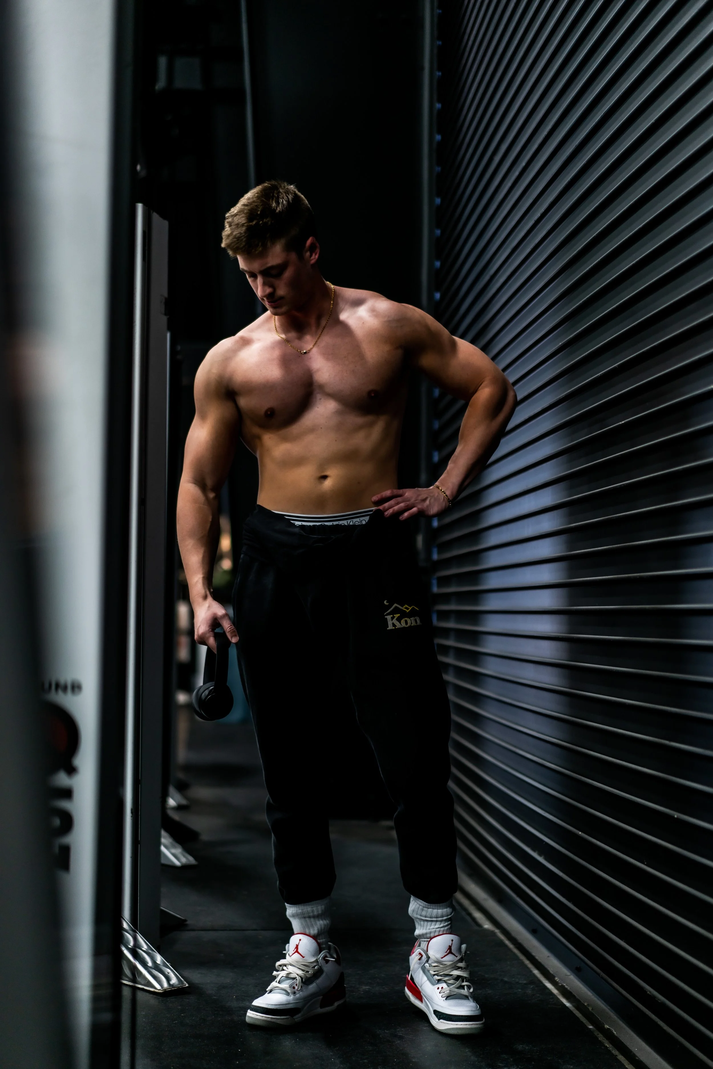 A shirtless young man with short brown hair stands in a dimly lit gym, holding a weight in his right hand. He wears black sweatpants with a gold word that looks like 'Kön', white socks, and white sneakers with red accents. He appears to be resting between exercises, leaning against a metallic wall panel.