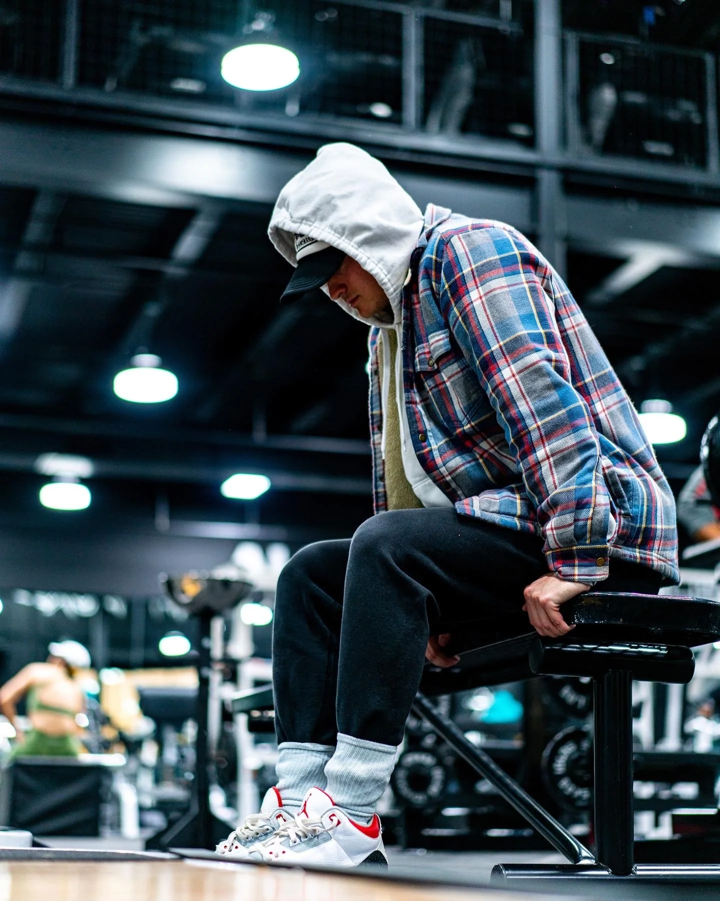 A man in a white hoodie and plaid shirt sitting on a bench in a gym, wearing sneakers with red accents.