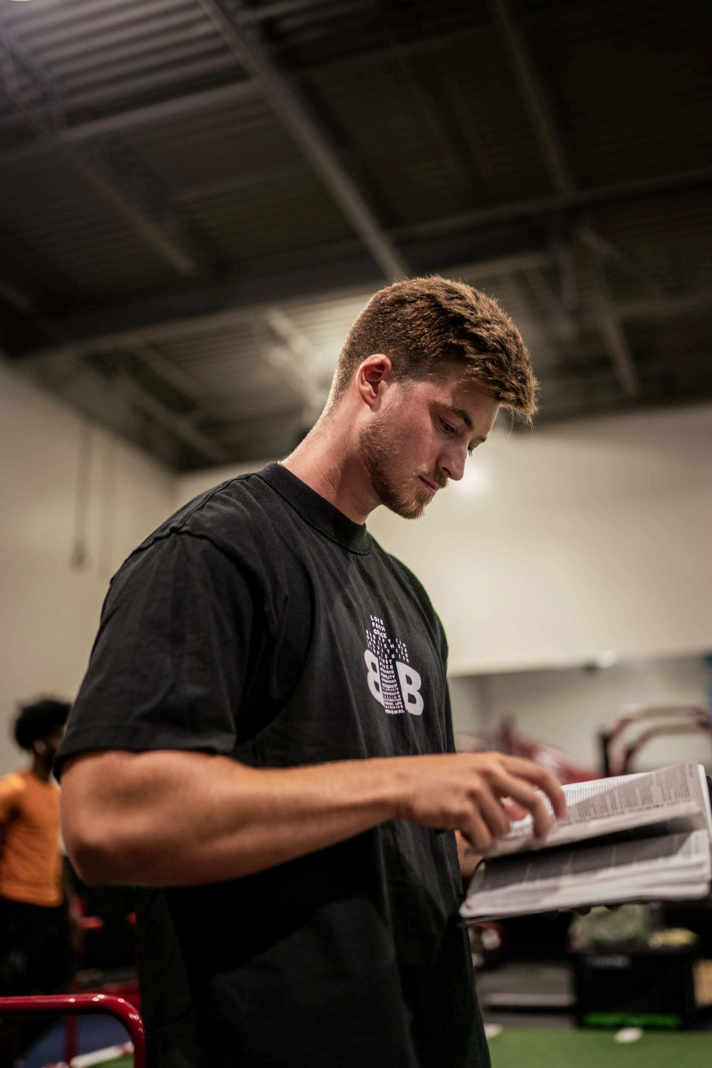 A young man with a beard and short brown hair, wearing a black T-shirt, is reading a book or magazine. He is in an indoor gym or training facility, with a high ceiling and other people in the background.