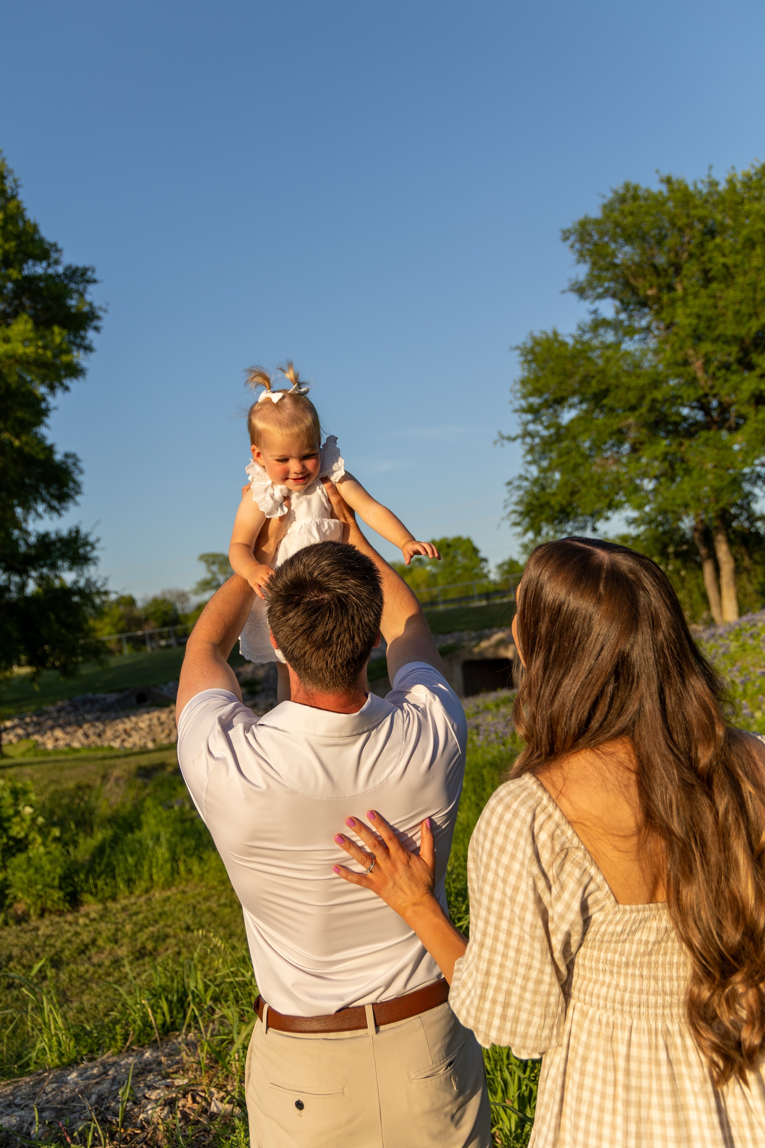 A man lifting a young girl during an outdoor daytime gathering, with a woman watching closely, against a background of trees and blue sky.