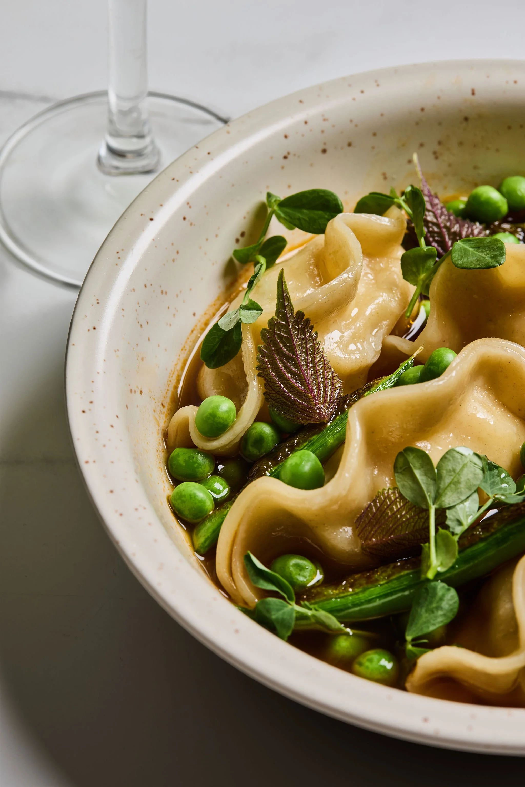 Close-up of a bowl of pasta with fresh herbs, peas, and broth, with a wine glass in the background.